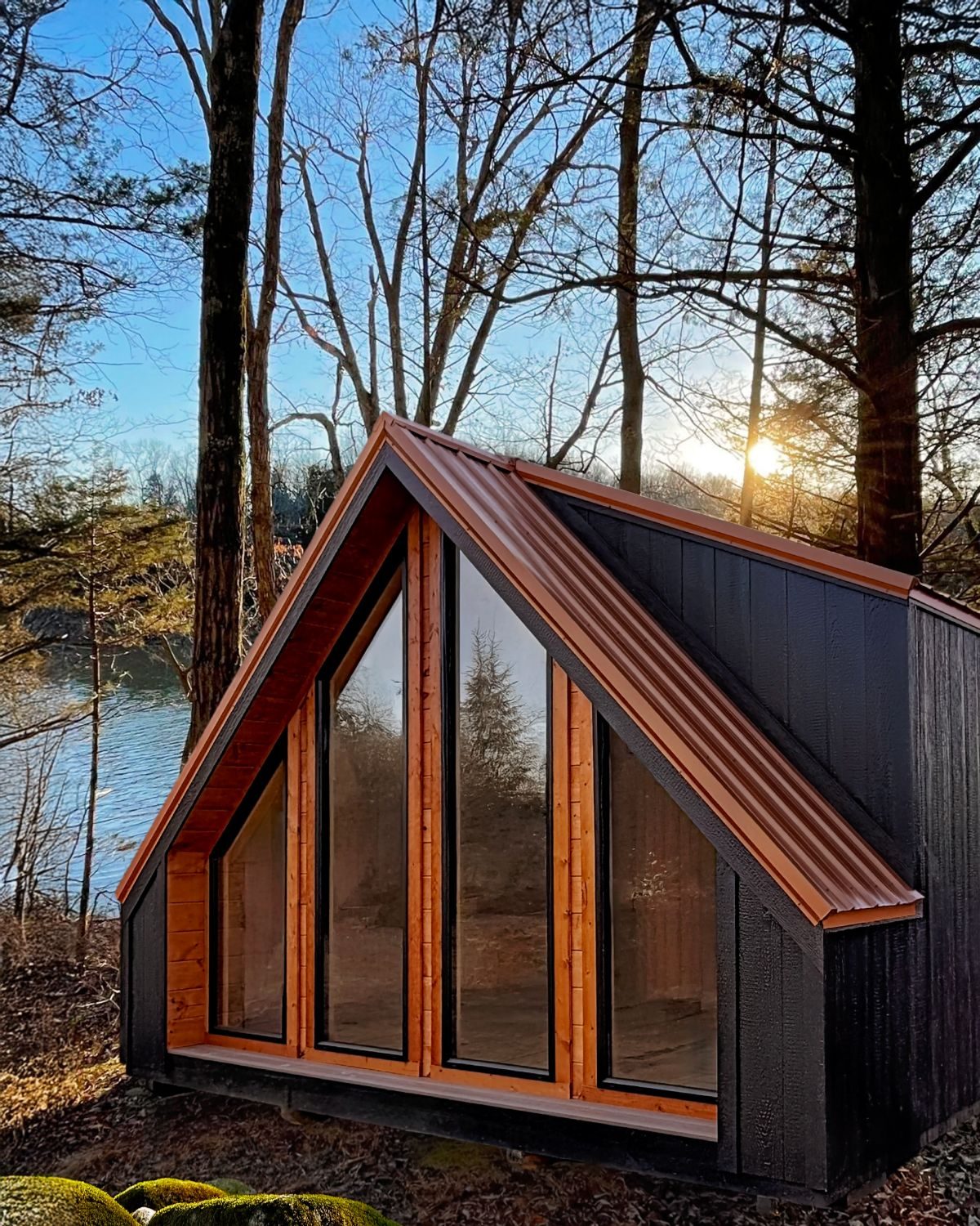 Black A-frame tiny home with a large glass gable window and copper-toned roof trim overlooking a lake