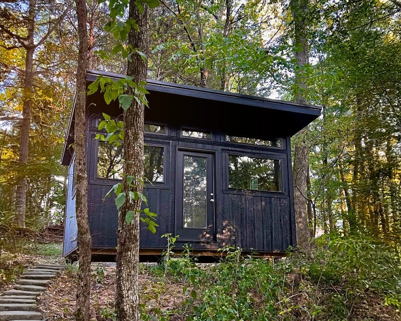 Black modern shed-roof tiny home with black door and windows, set in a forest