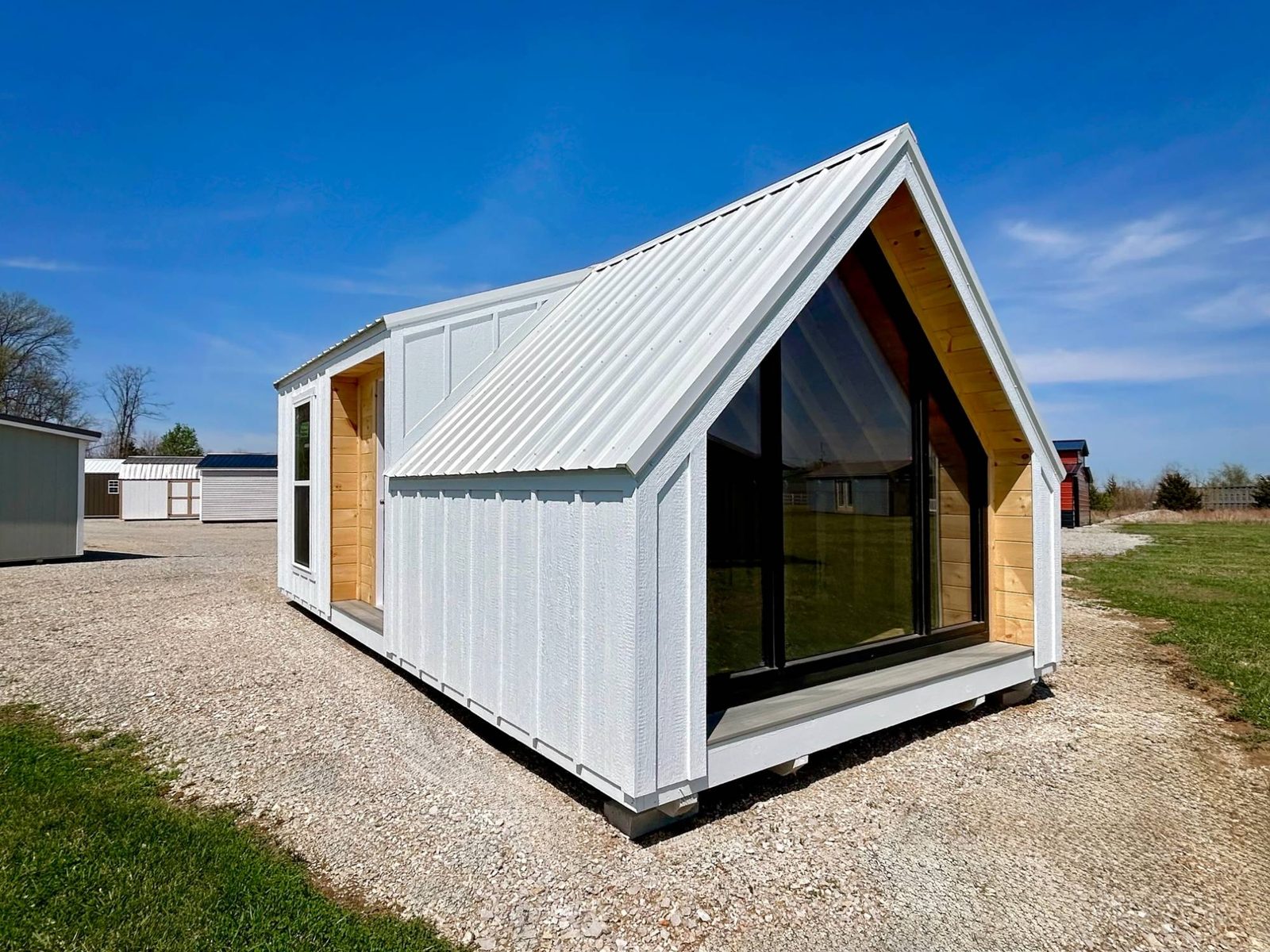 White A-frame tiny home with a covered porch, a wood door, and a large black-trimmed triangular window
