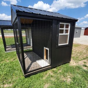 Black metal dog kennel with wire mesh sides and a corrugated metal roof, set on a grassy yard against a blue sky with clouds.