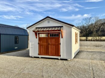 White shed with red double doors and a small metal awning, set on a gravel lot under a bright blue sky.