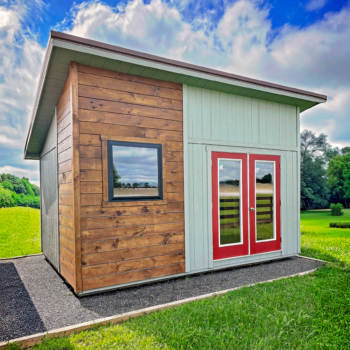 Small wooden shed with green siding and red double doors on a paved path, sunny grassy field in the background.