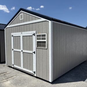 Gray storage shed with white trim and double doors on a gravel lot, under a clear blue sky with a few clouds