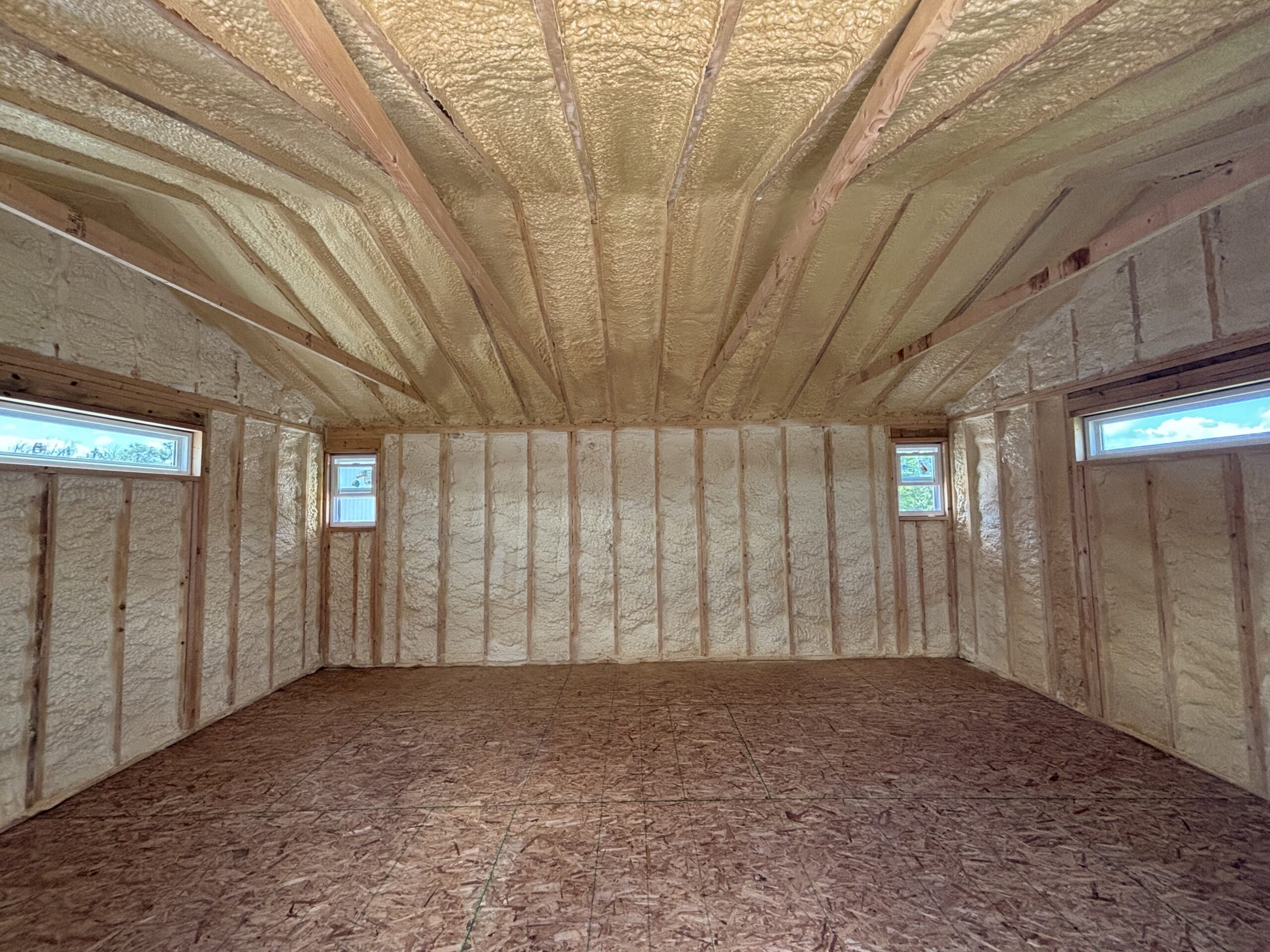 Interior of an unfinished room with spray-foam insulation on walls and ceiling, exposed wooden studs, and a plywood floor.