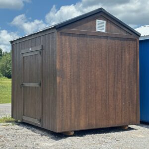 Brown wooden shed on a gravel lot, with a single tall door and small vent near the roof, placed beside a blue building outdoors.