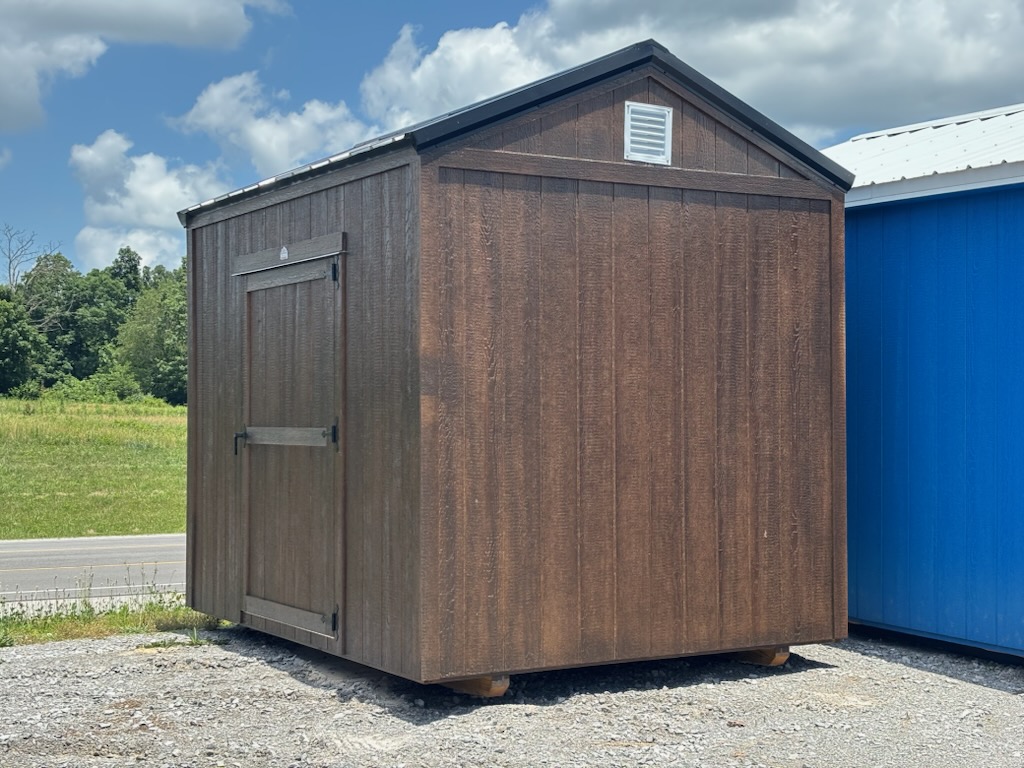 Brown wooden shed on a gravel lot, with a single tall door and small vent near the roof, placed beside a blue building outdoors.