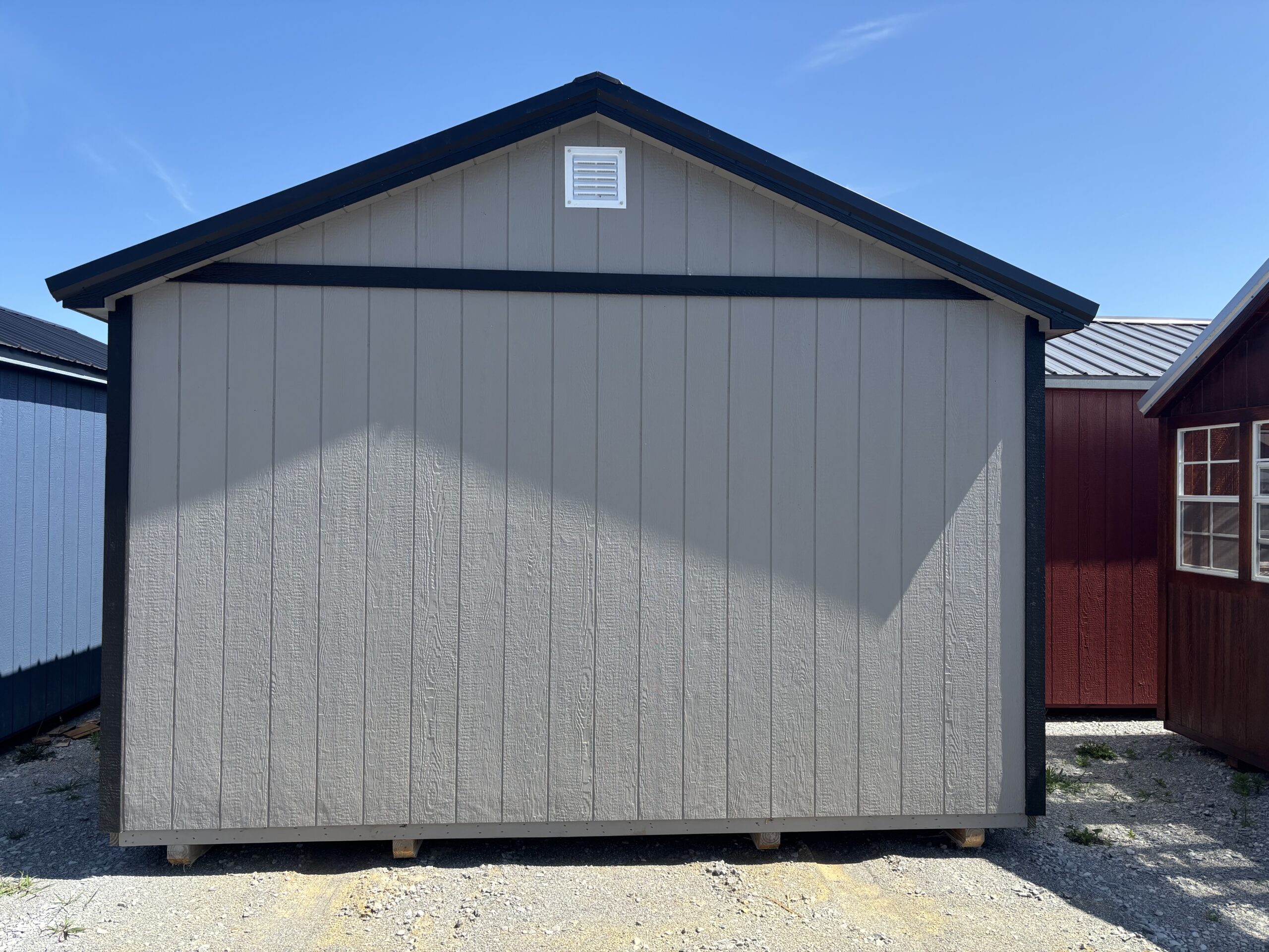 Front view of a light gray shed with dark blue trim and a small white vent near the roof peak on a gravel lot; a red shed is visible to the right.