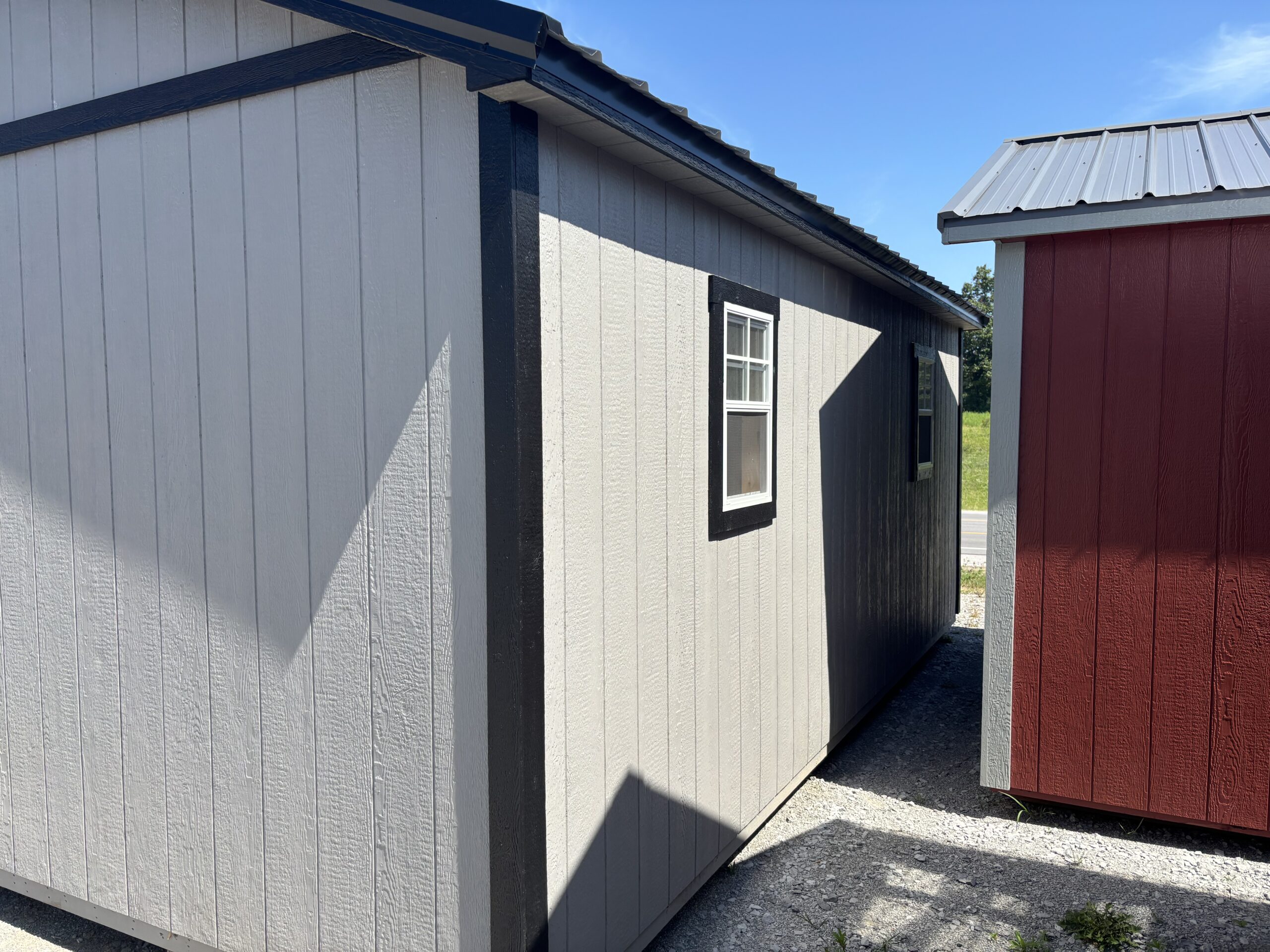 Two small wooden sheds with gray siding and black trim sit side by side in a sunny alley, with a red shed nearby and metal roofs above.