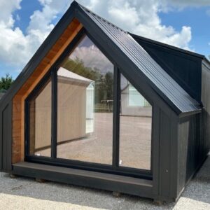 Black A-frame shed with a large front glass wall and wooden interior on a gravel lot, under a blue sky with clouds.