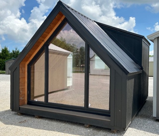 Black A-frame shed with a large front glass wall and wooden interior on a gravel lot, under a blue sky with clouds.