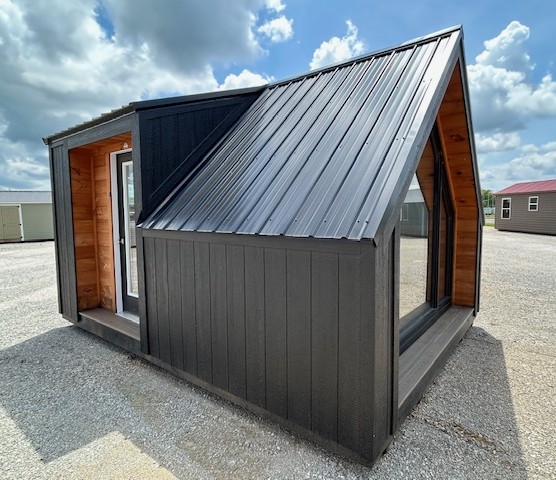 Small black metal cabin with a steep sloped roof, open doorway showing a wooden interior, on a gravel lot under a blue sky.