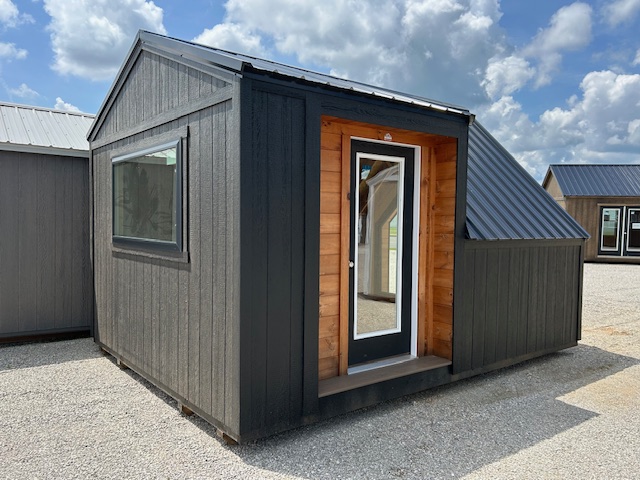 Dark gray modular shed with a small wooden-framed entry door and a side window on a gravel lot