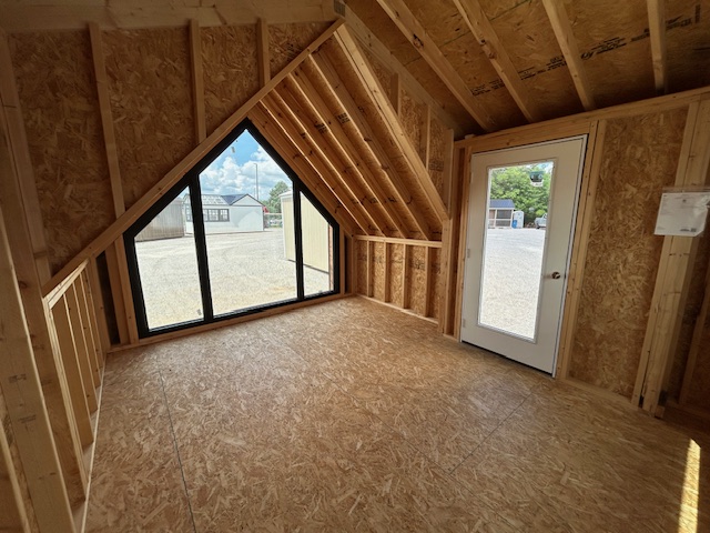 Unfinished attic room with exposed wooden framing and plywood walls; a large triangular window and a glass exterior door lead outside.