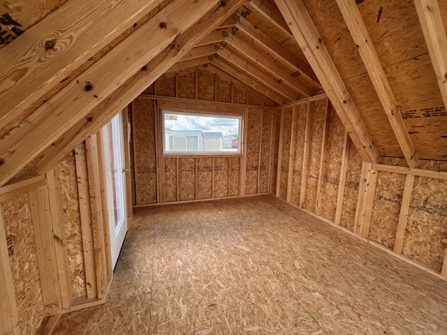 Unfinished attic with exposed wooden framing, plywood floor, and sloped rafters; window at far wall shows houses outside.