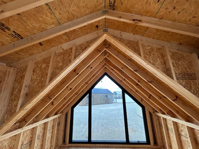 Interior of an unfinished attic with exposed wooden rafters and OSB sheathing, featuring a large triangular dormer window overlooking a gravel area and buildings outside.