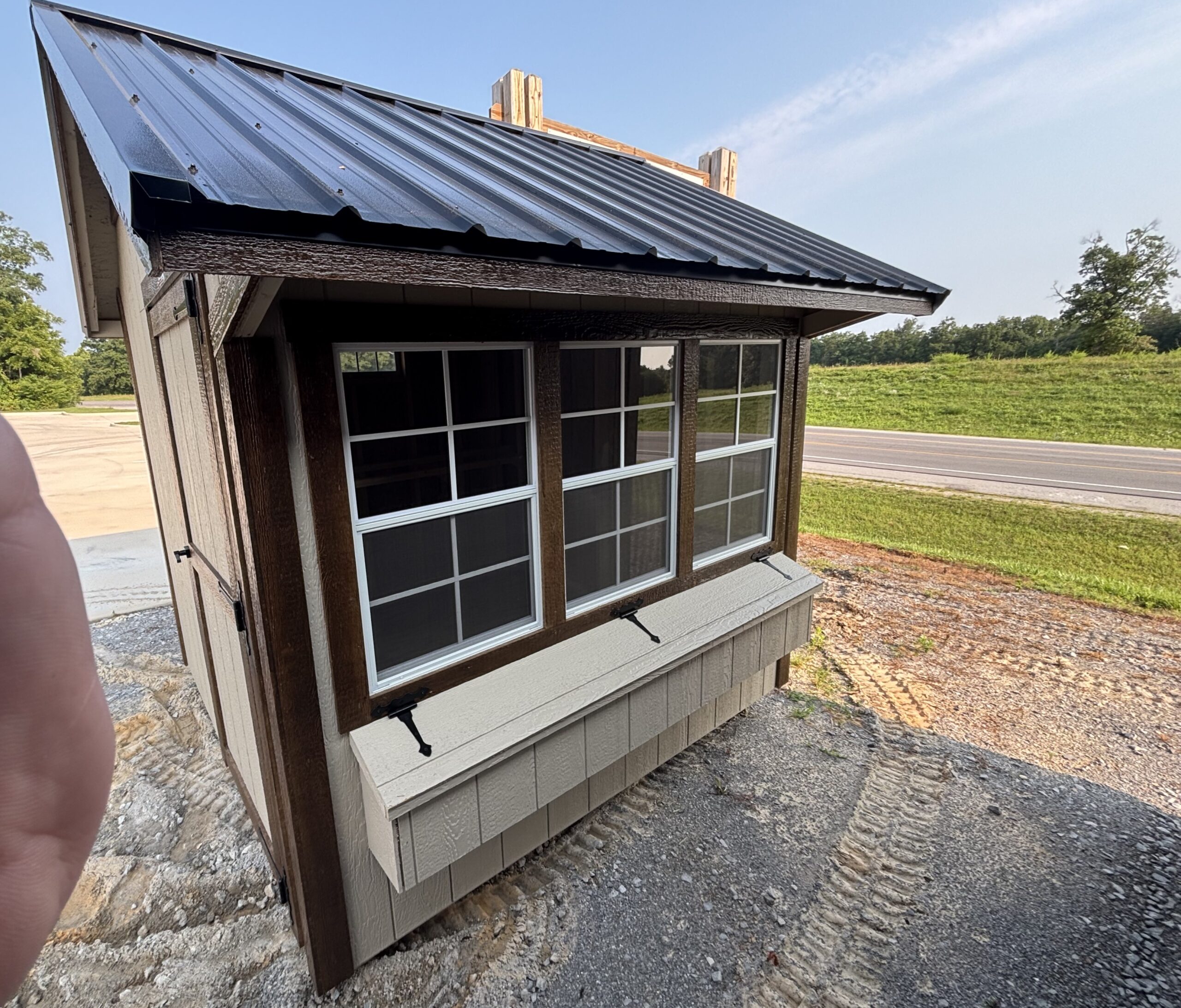 Close-up of a small wooden building with a sloped metal roof, large front window, and dark brown trim, near a road in a grassy area. A finger is visible in the lower-left corner.