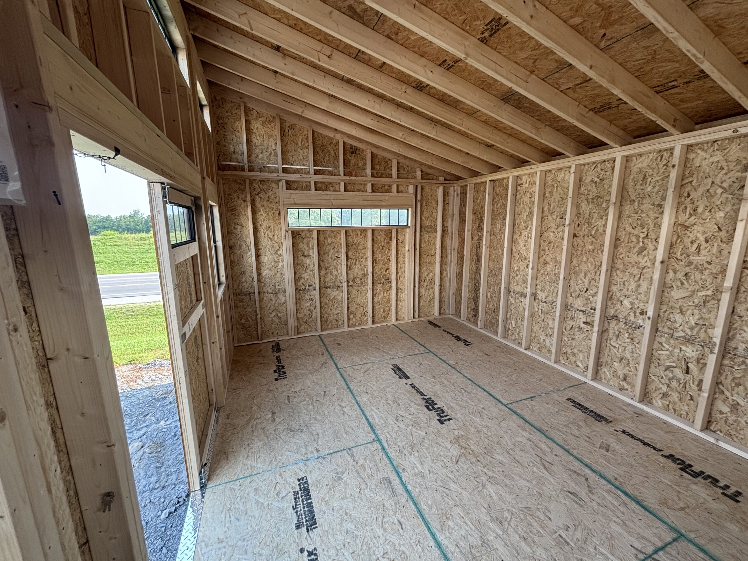 Interior of an unfinished, wood-framed room with exposed studs and roof trusses; a small window lets in daylight from outside.