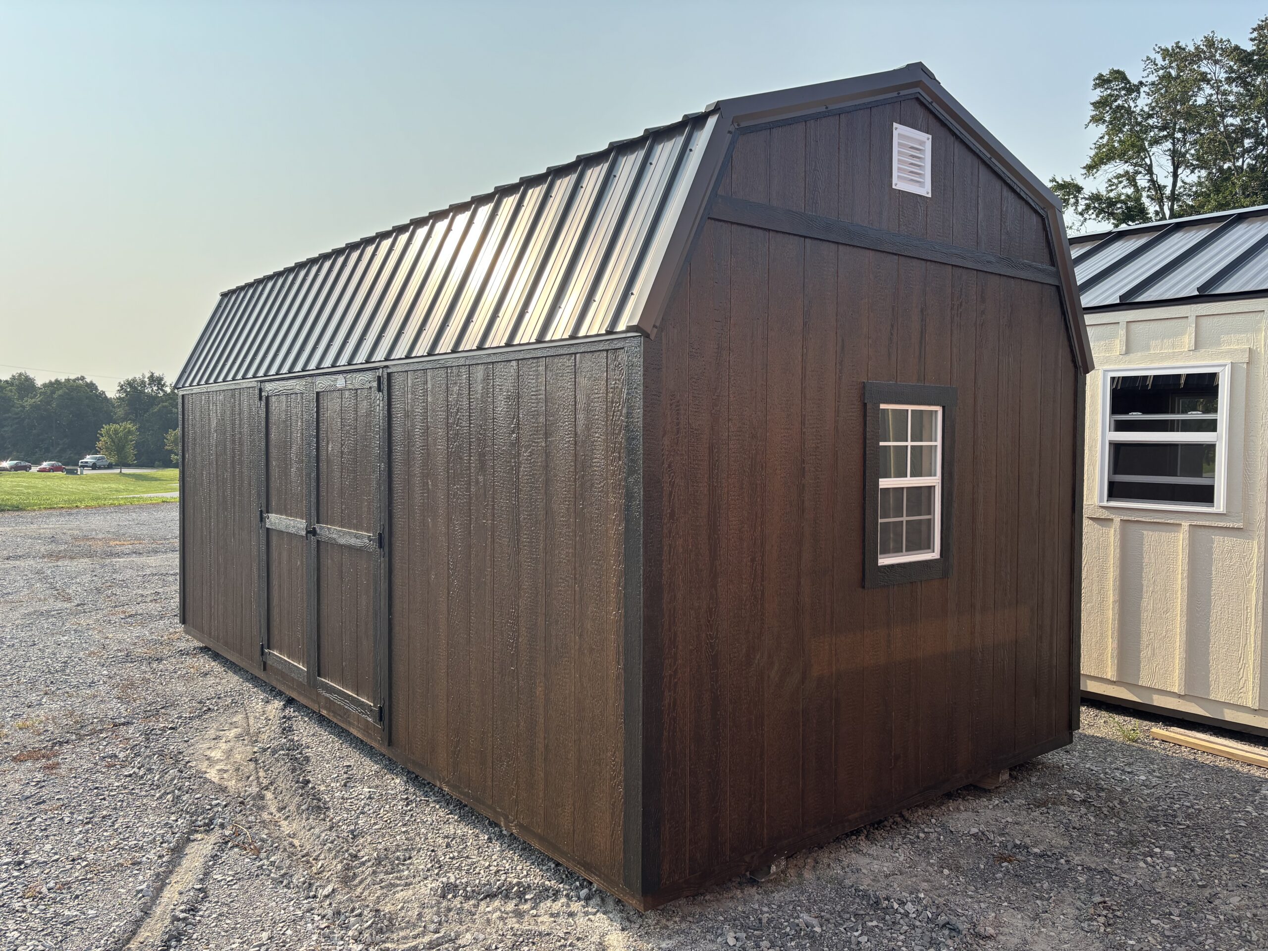 Brown wooden storage shed with a metal gable roof and small window on a gravel lot.