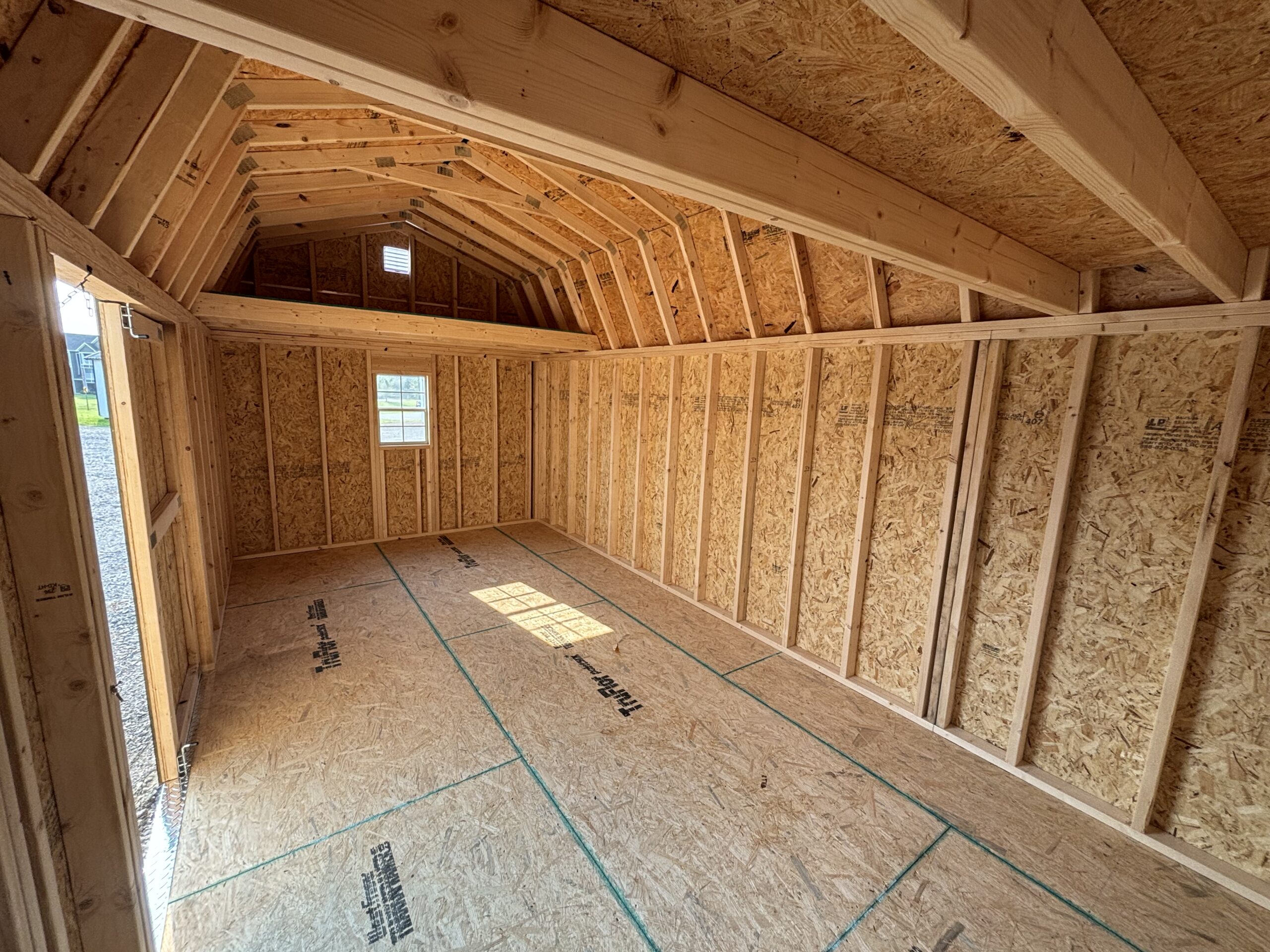 Unfinished attic with exposed framing, plywood walls, and angled roof trusses, a window at the far wall letting in light.