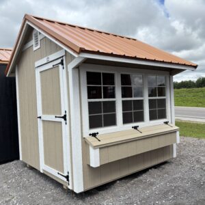Beige wooden shed with white trim, large windows, and an orange metal roof on a gravel lot under a cloudy sky.