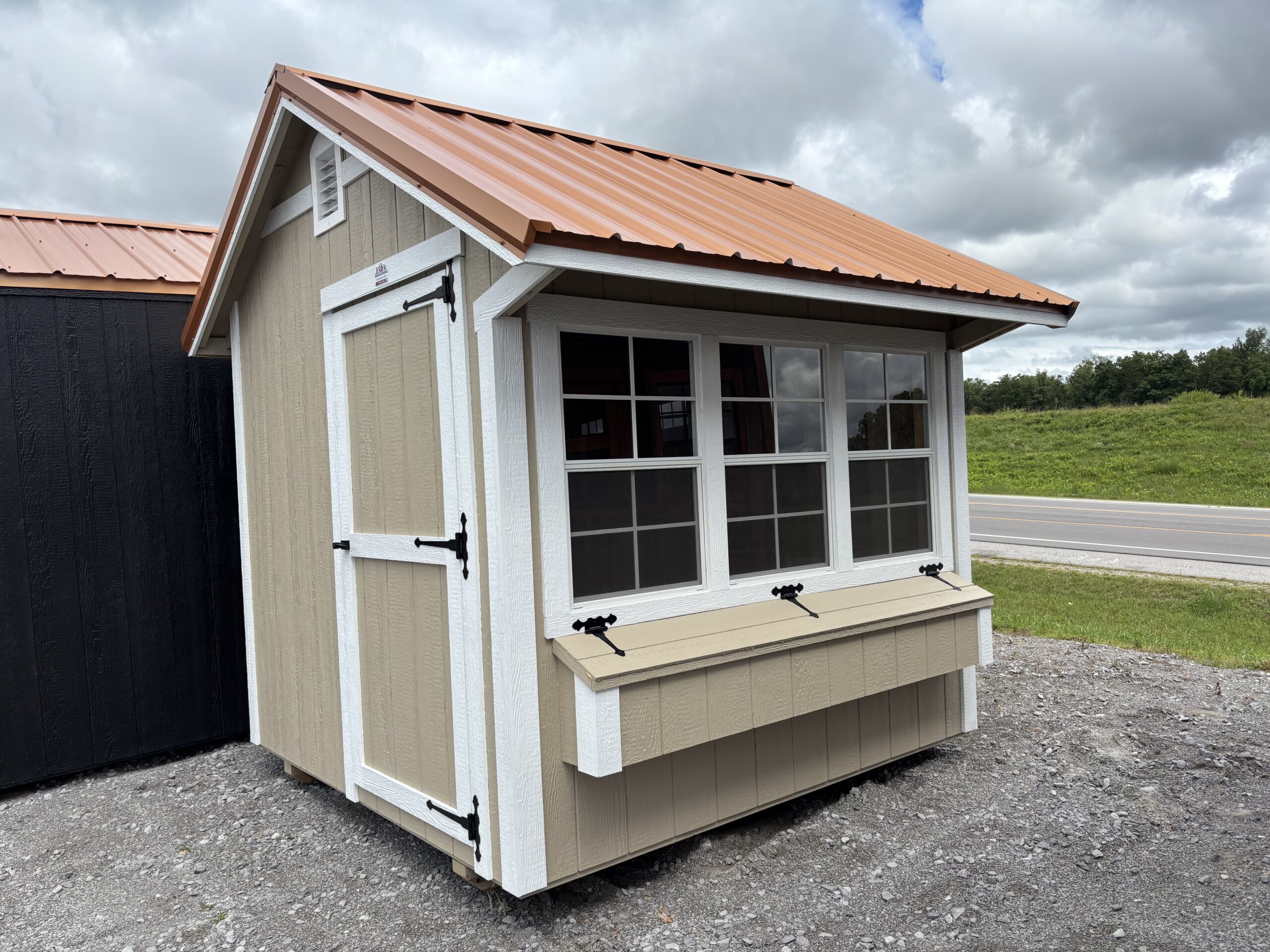 Beige wooden shed with white trim, large windows, and an orange metal roof on a gravel lot under a cloudy sky.