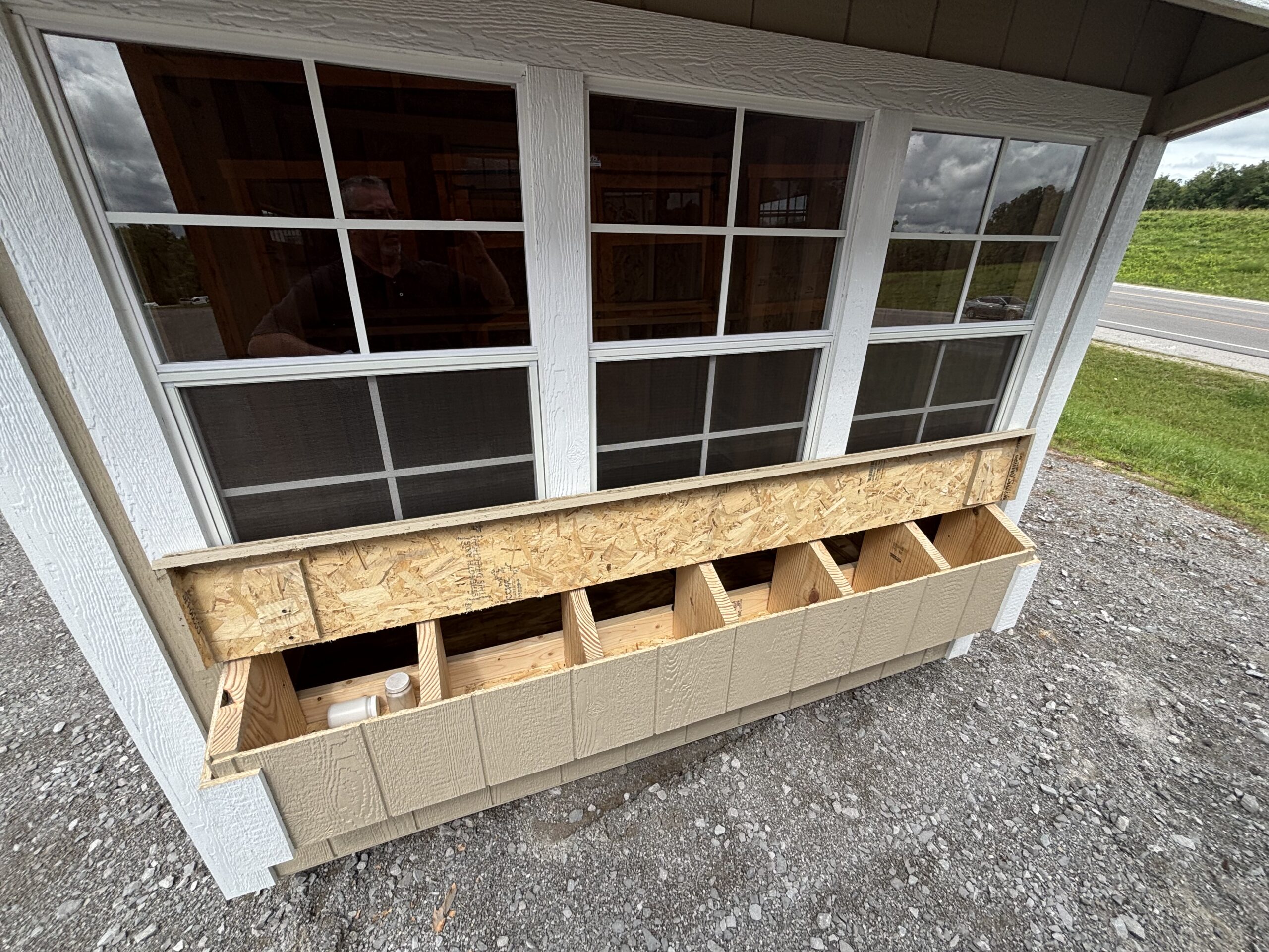 Exterior window of a house under construction with white trim, plywood formwork, and stacked lumber nearby on a gravel surface.