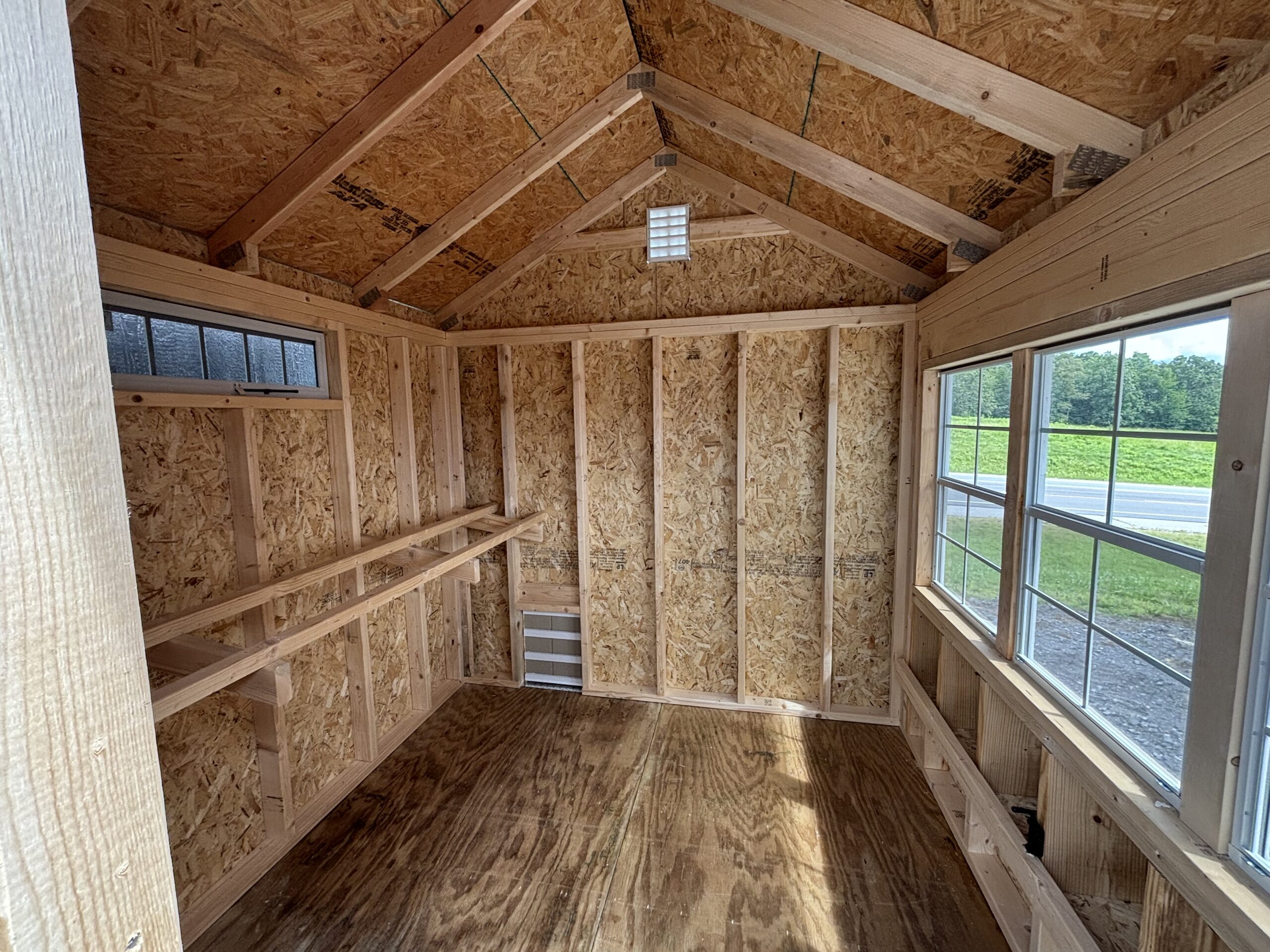 Interior of a small unfinished wooden shed with OSB walls, exposed studs, and built-in shelves along both walls, plus a window and vent near the peak.