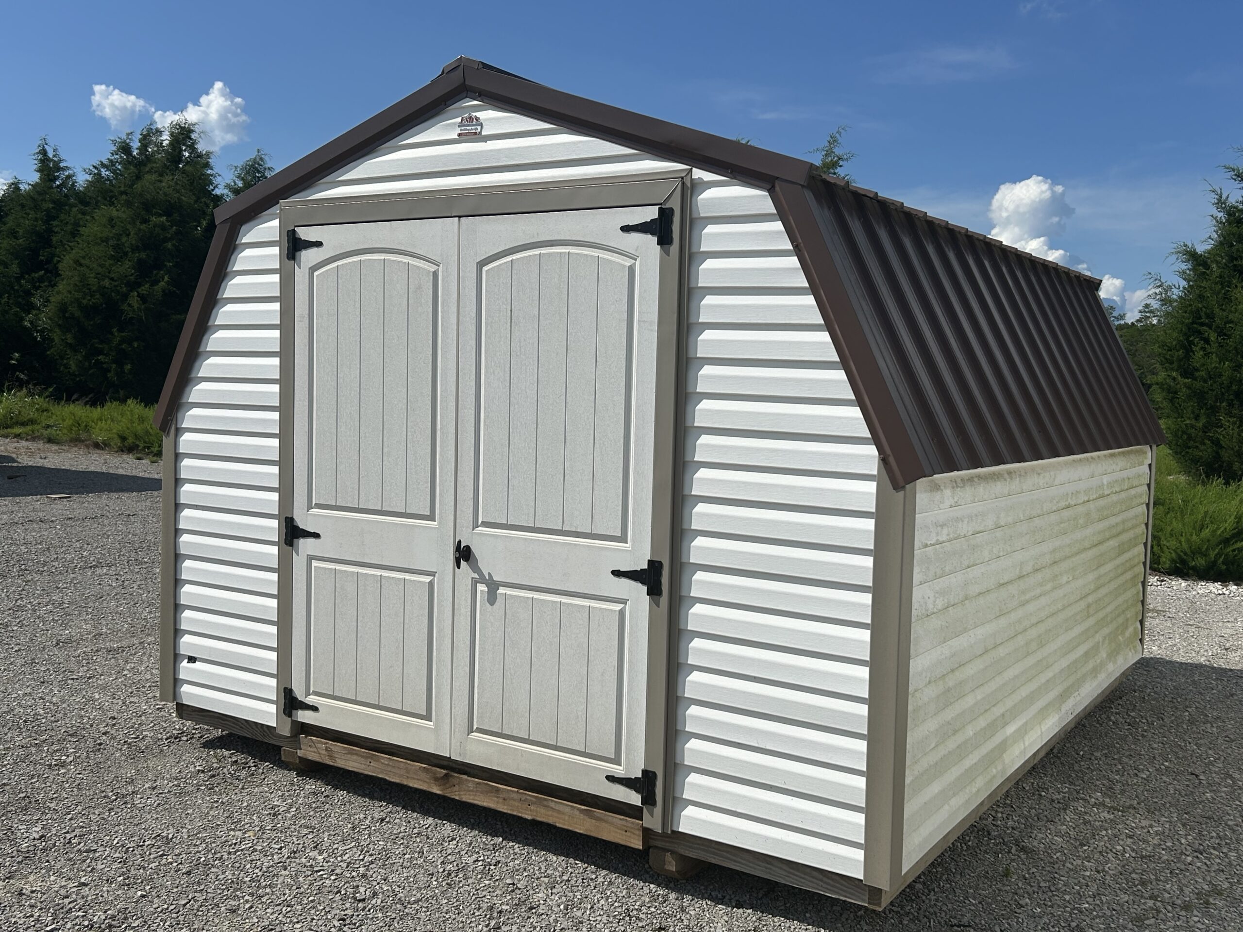 White outdoor storage shed with double arched doors, white siding, and a brown peaked roof on a gravel lot.