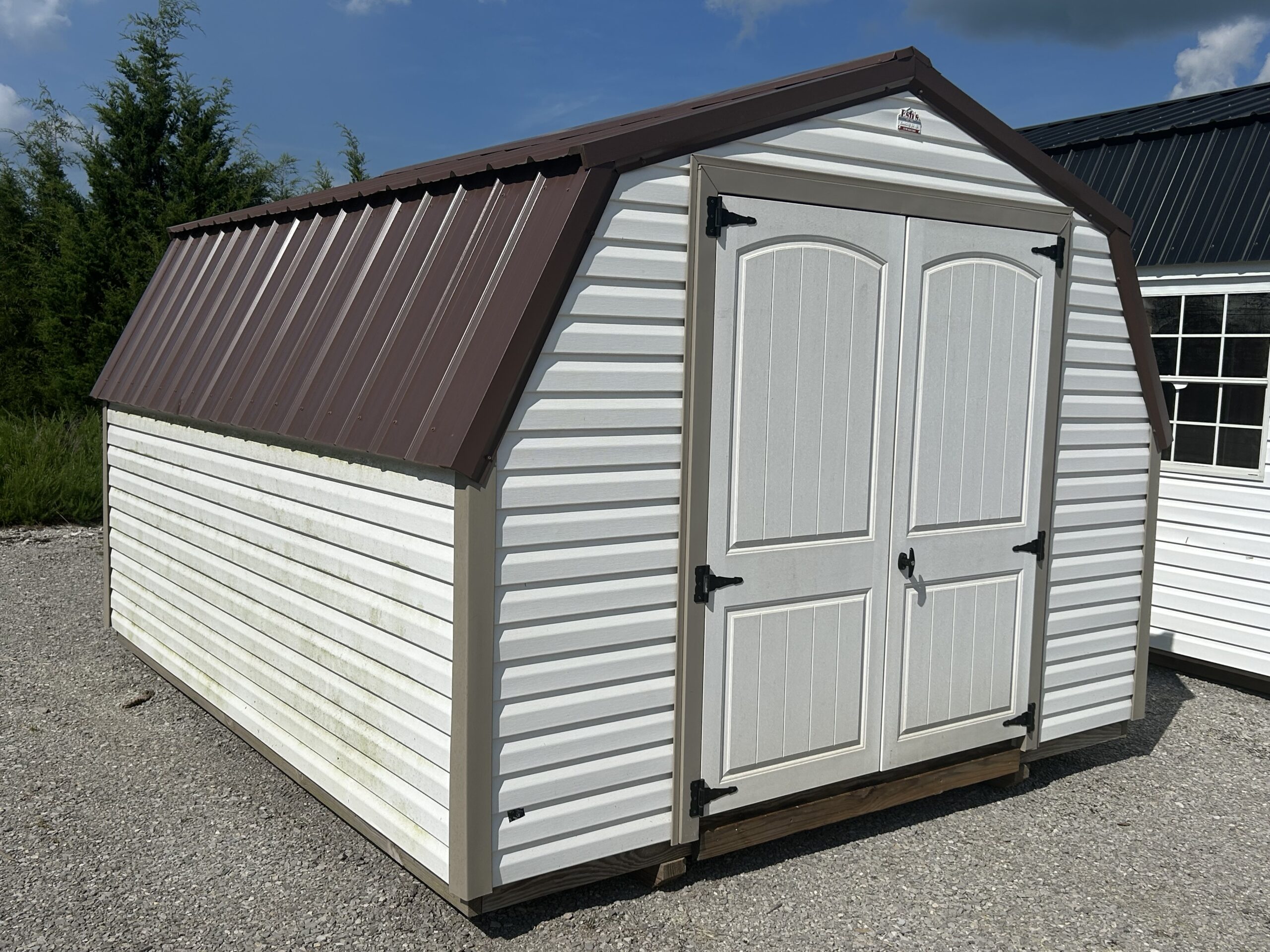 White storage shed with a brown metal roof and double paneled doors on a gravel lot, outdoors.