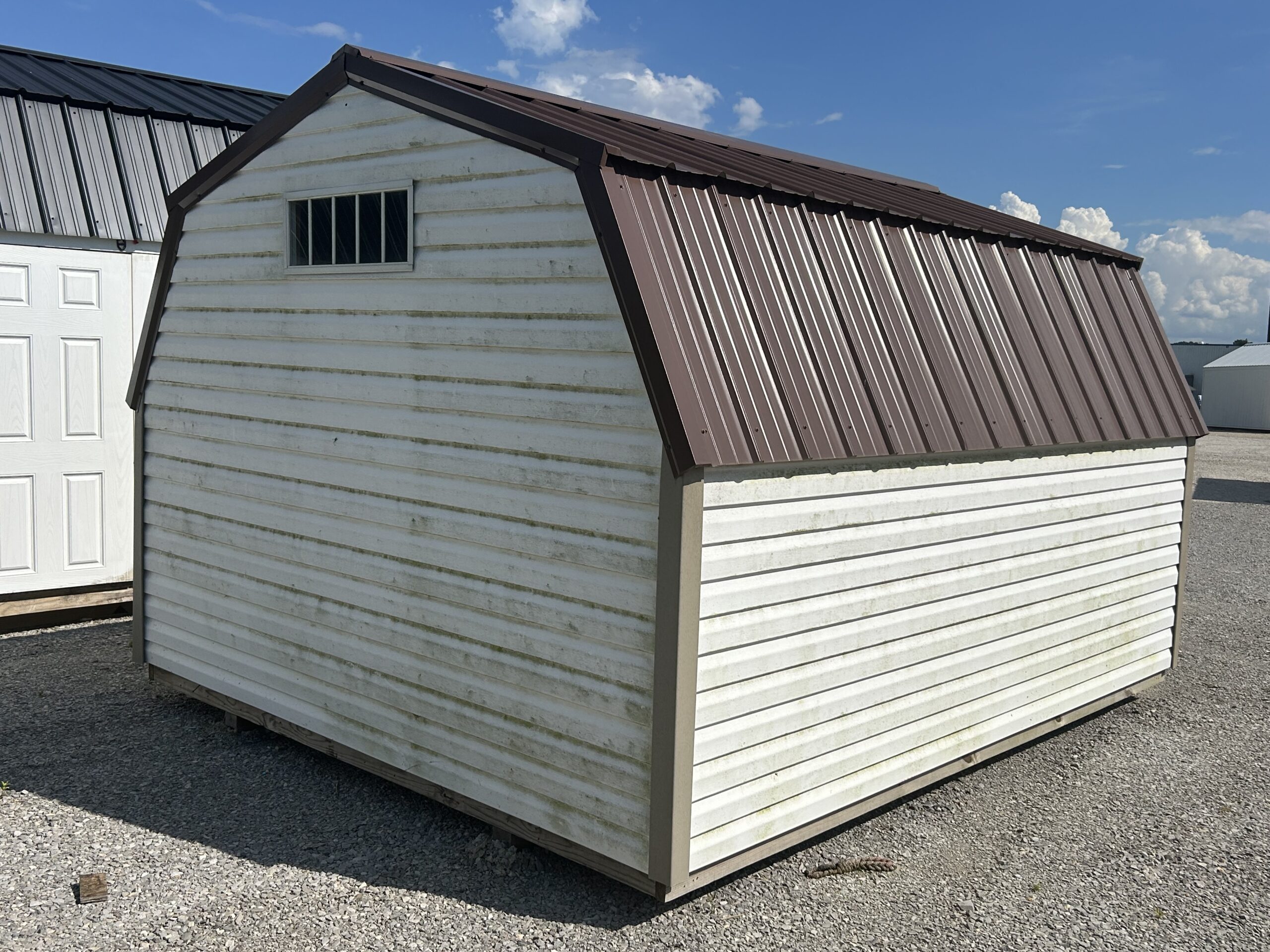 White weathered storage shed with a brown metal gabled roof on a gravel lot under a blue sky, next to another shed with white doors.
