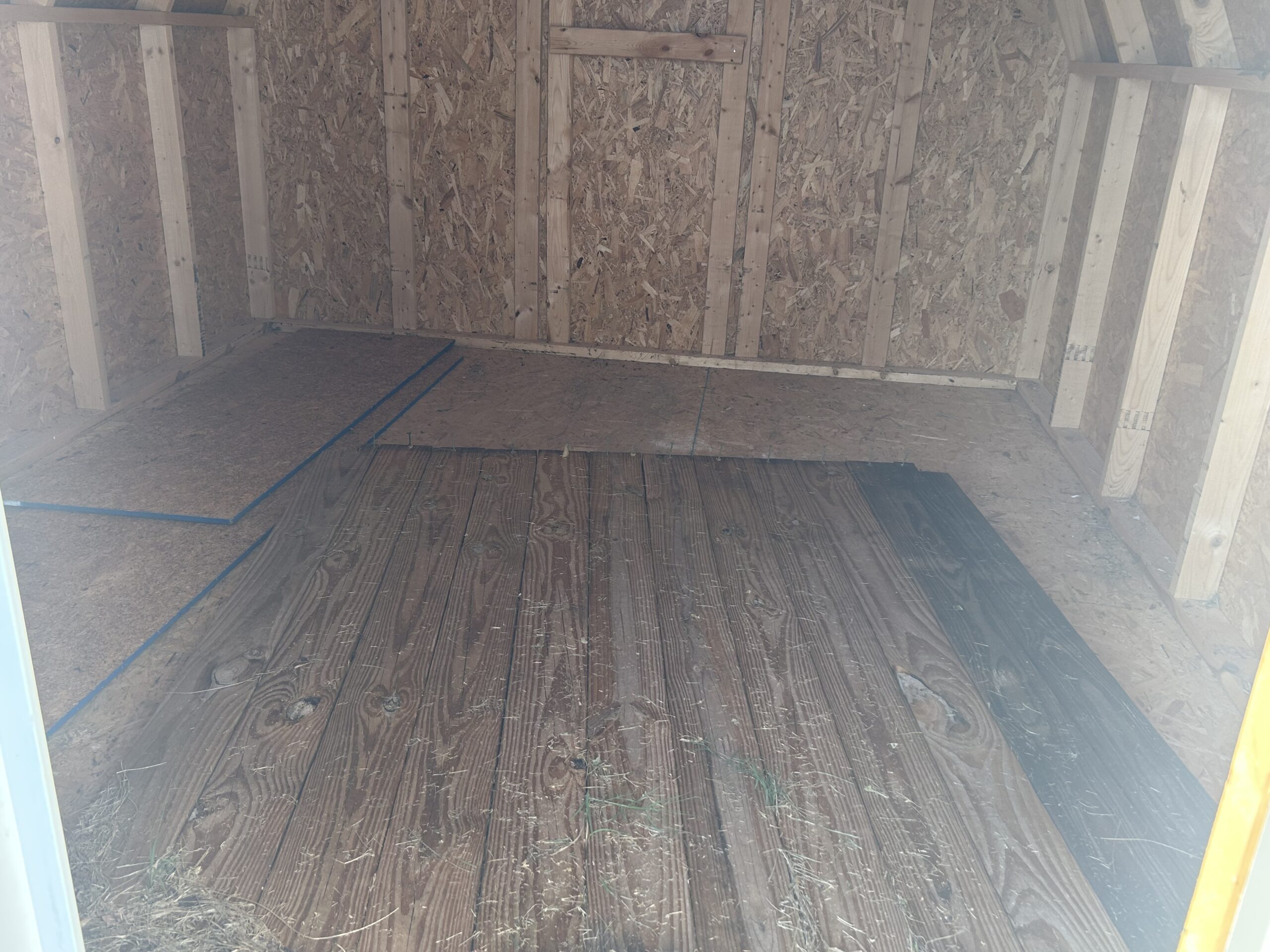 Interior of an unfinished wooden attic with OSB walls, vertical studs, and taped outlines on the floor marking a area to build.