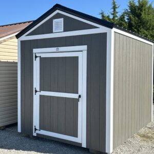 Gray garden shed with white trim and a small vent under the peaked roof, located on a gravel surface with another beige shed nearby and evergreen trees in the background.