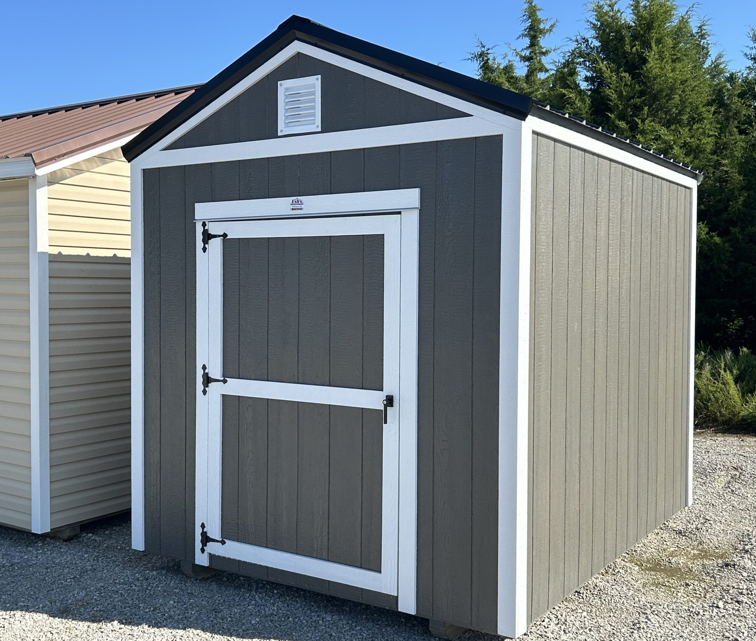 Gray garden shed with white trim and a small vent under the peaked roof, located on a gravel surface with another beige shed nearby and evergreen trees in the background.