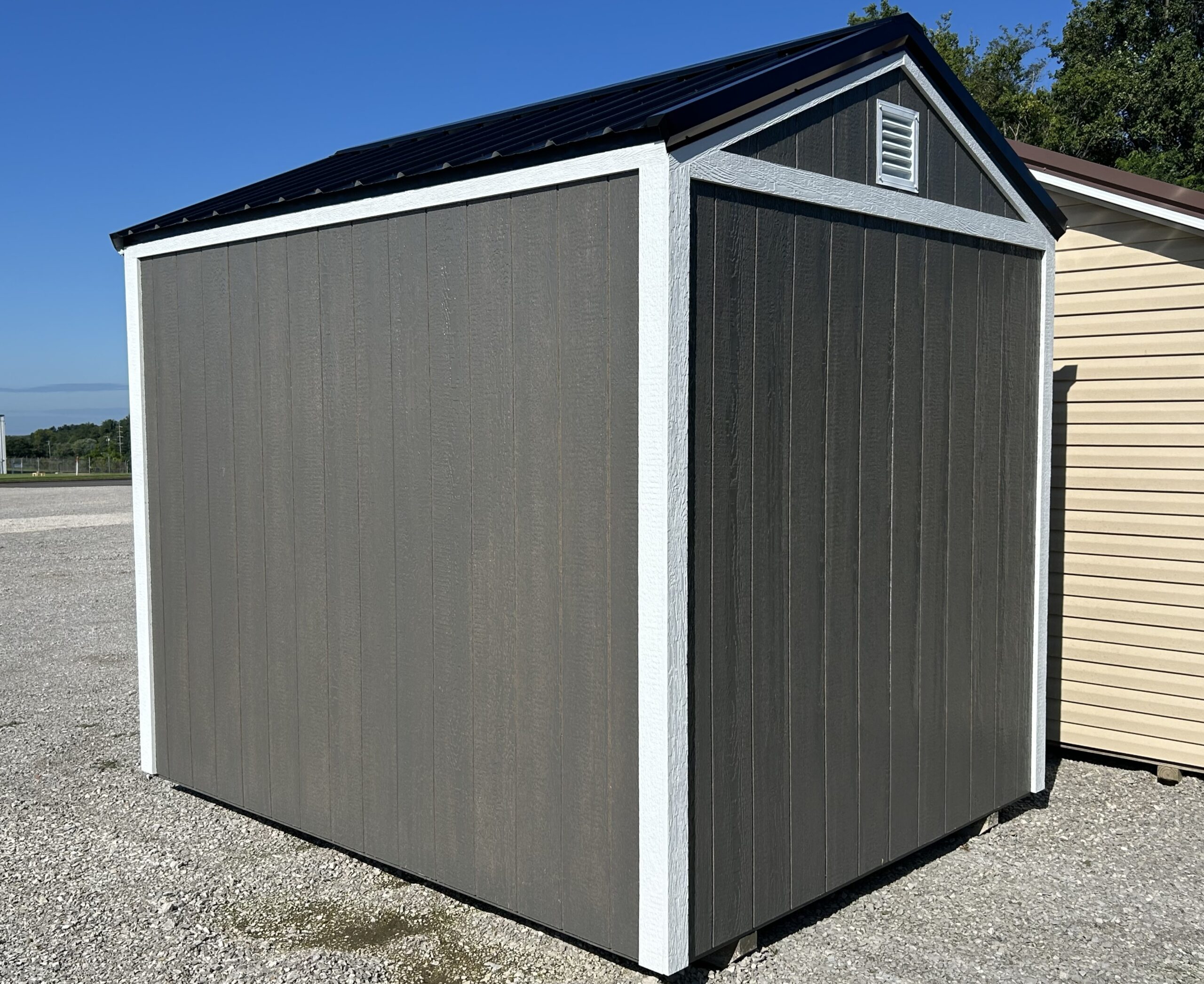 Grey outdoor storage shed with white trim and a vented gable, set on a gravel lot under a clear blue sky.
