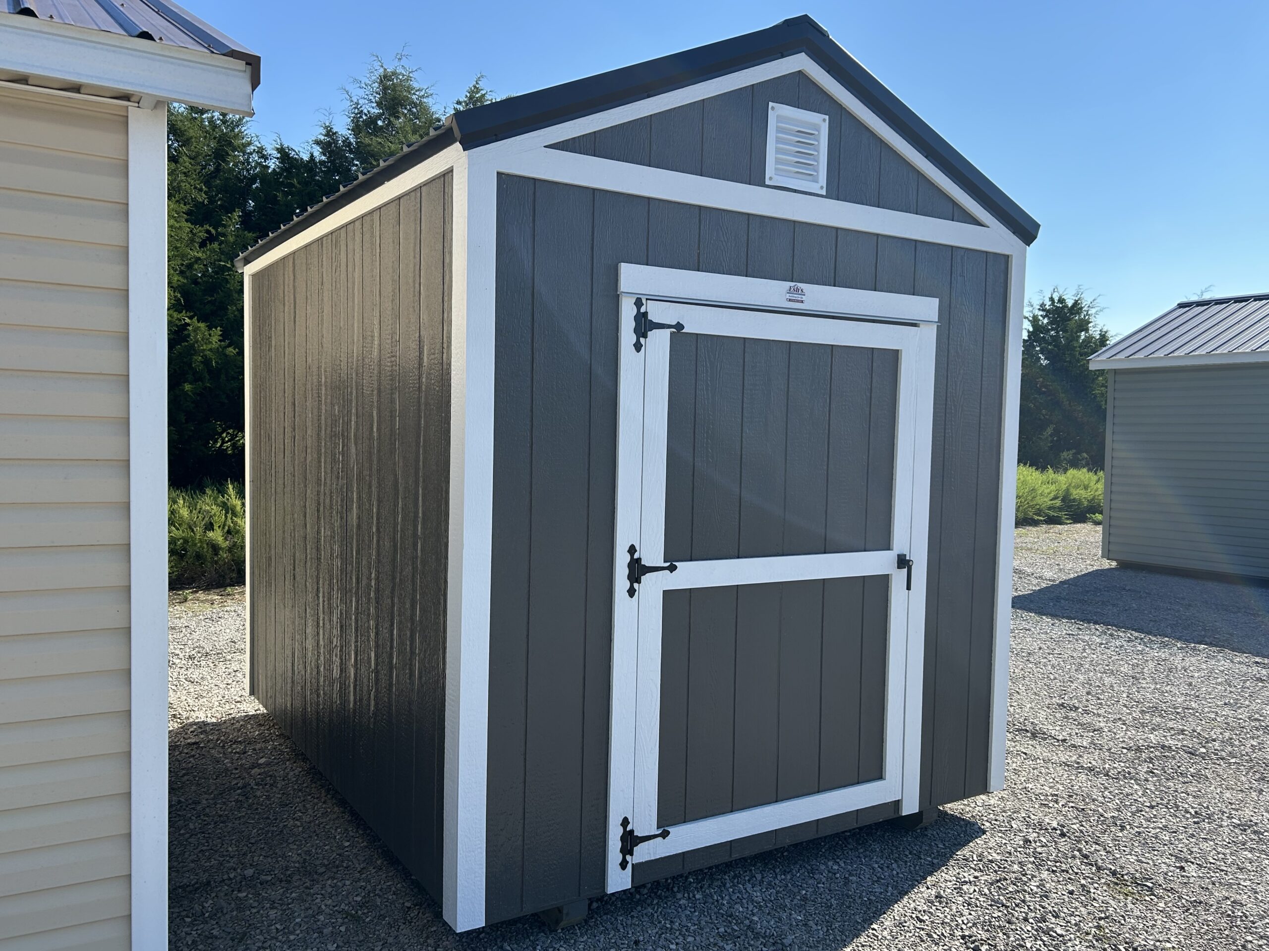 Gray wooden storage shed with white trim and two black decorative hinges on a barn-style double door, standing on a gravel lot under a clear blue sky.