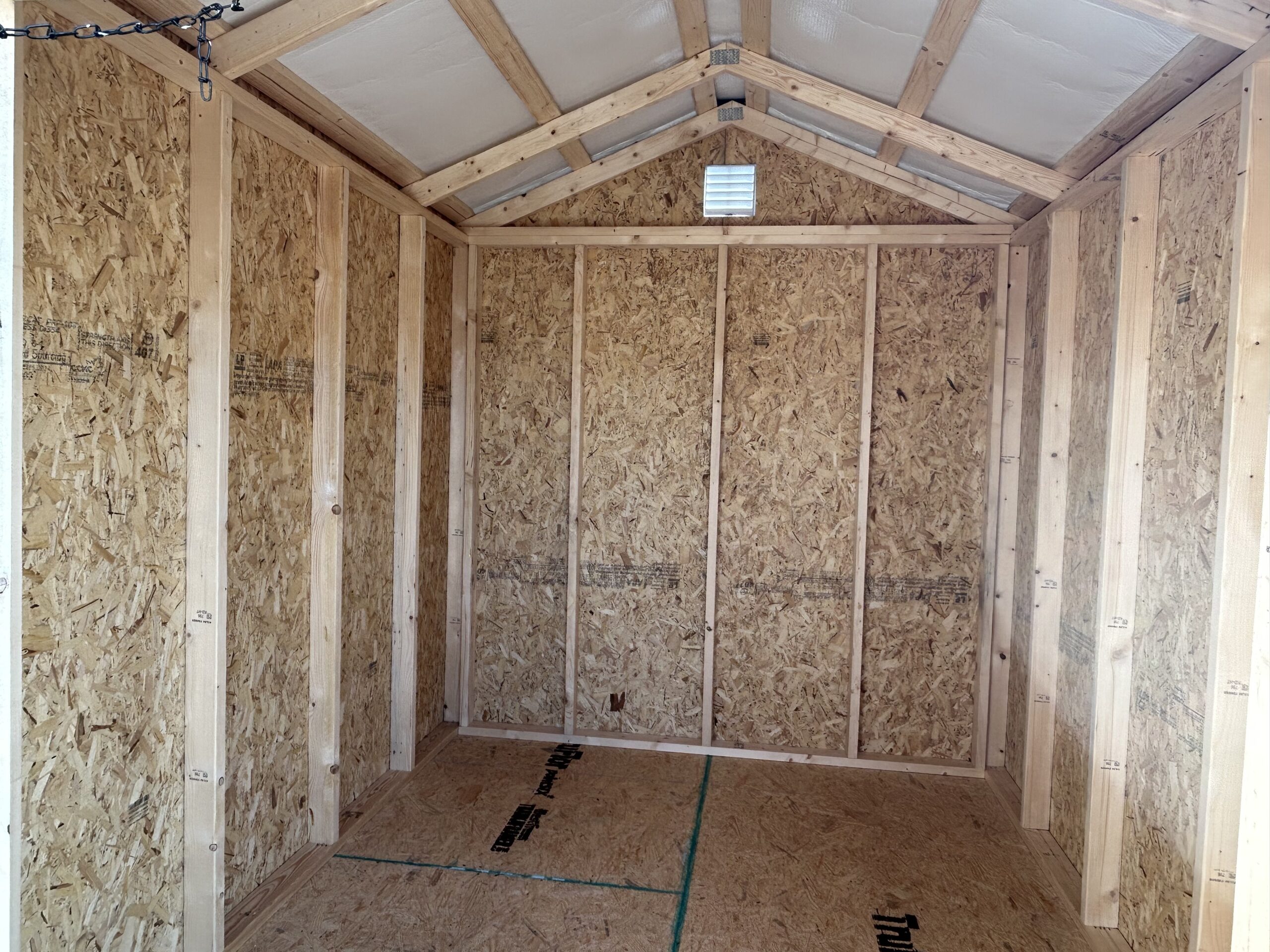 Interior of an unfinished shed with exposed framing, oriented strand board walls, and triangular roof trusses; a small vent is on the back wall and a chain hangs from the ceiling on the left side.
