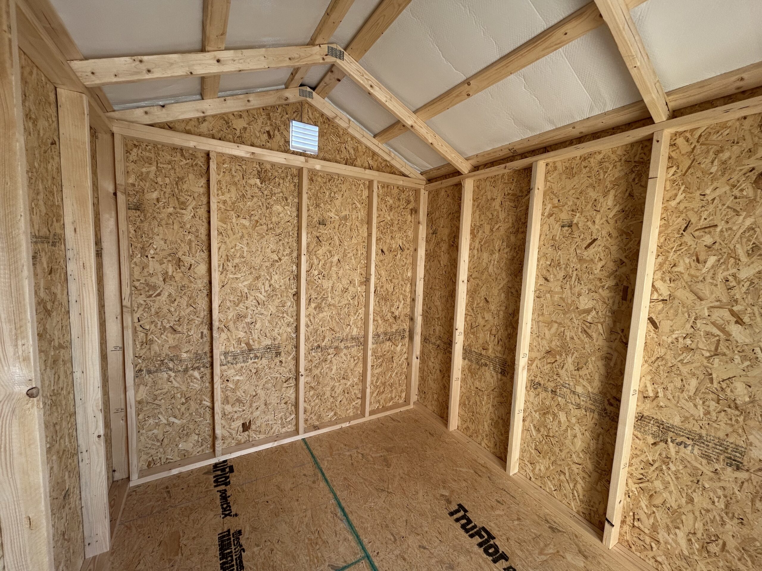 Interior view of a wooden framed room under construction with OSB walls, vertical studs, and a pitched ceiling with exposed rafters and a small wall vent.