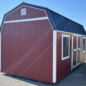 Red wooden shed with white trim and a black metal roof, set on a gravel lot under a clear blue sky.