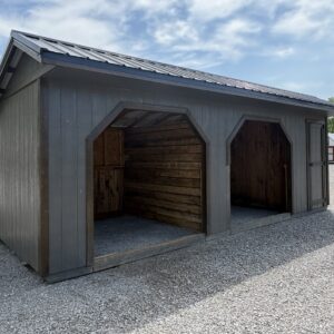Gray wooden storage shed with two large arched openings and a side door on a gravel lot under a blue sky.