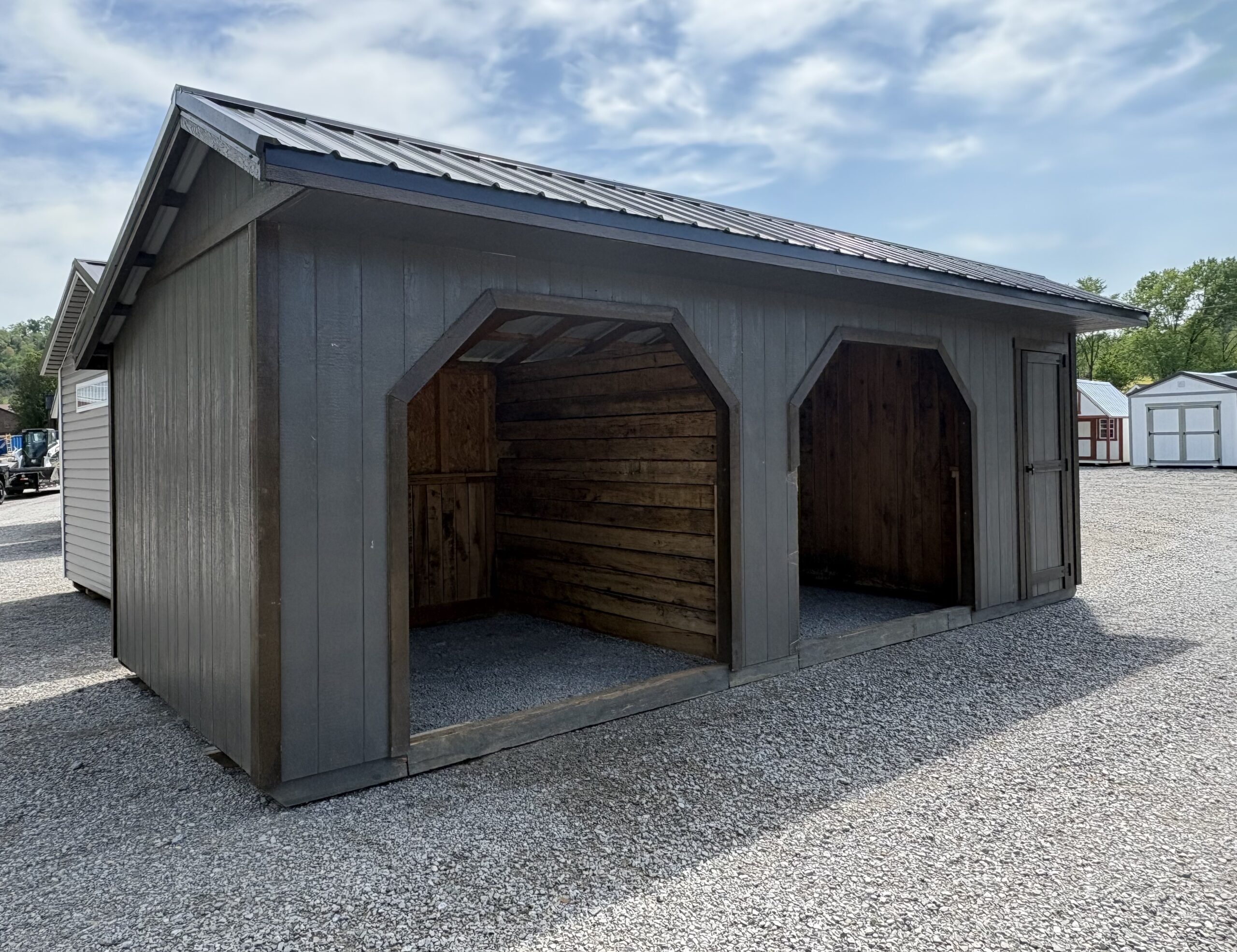 Gray wooden storage shed with two large arched openings and a side door on a gravel lot under a blue sky.