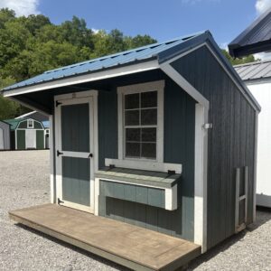 Small blue shed with a wire-menced window, a wooden ramp, and a metal roof against a gravel lot and forested hillside.