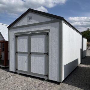 White wooden shed with double barn doors, gray trim, and a vent, set on a gravel lot under a partly cloudy blue sky