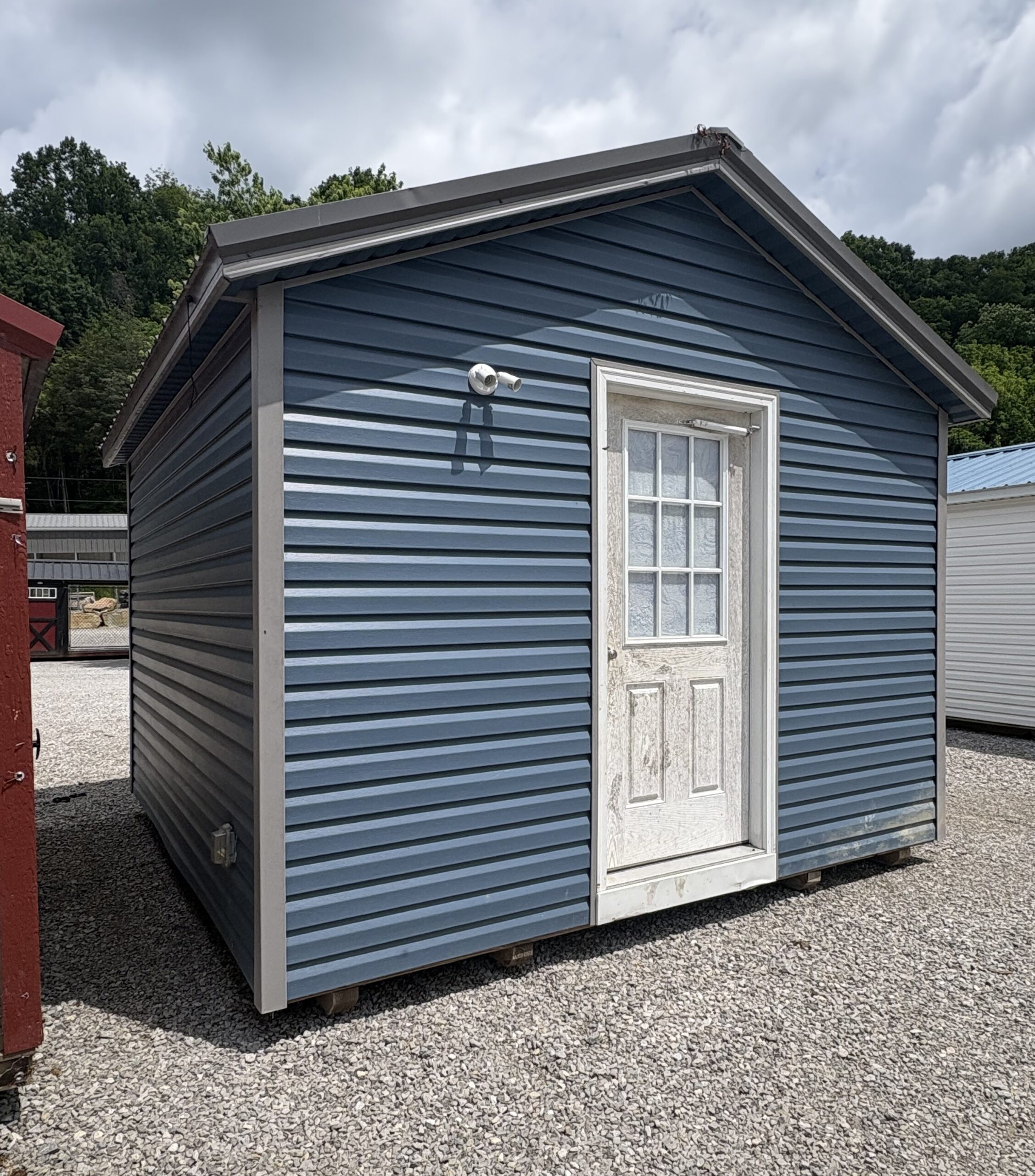 Blue metal siding storage shed with a white framed door and window on a gravel lot under a cloudy sky.