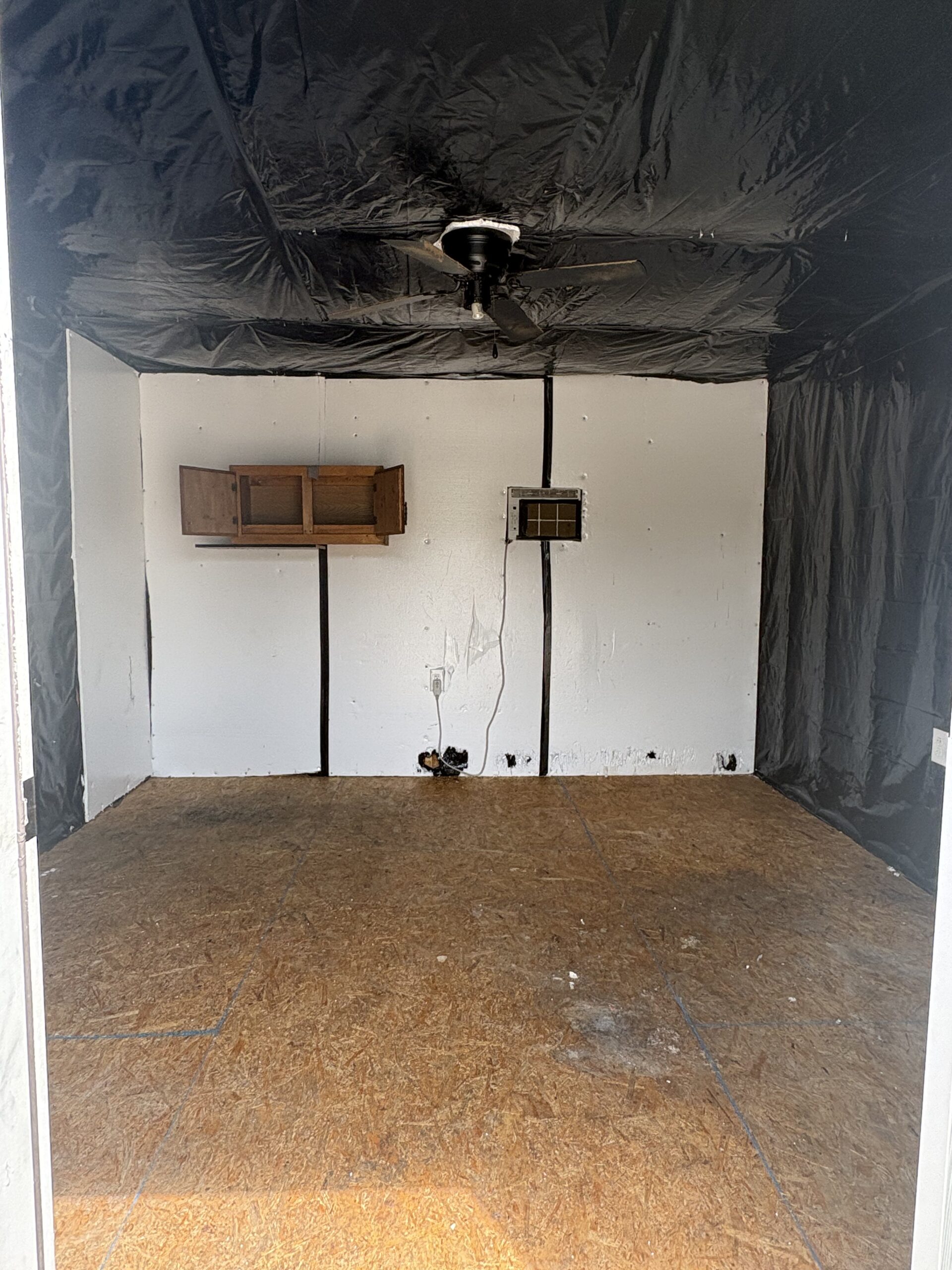 Empty room with black plastic walls, a ceiling fan, and a wall-mounted wooden cabinet on the back wall with a small vent nearby.
