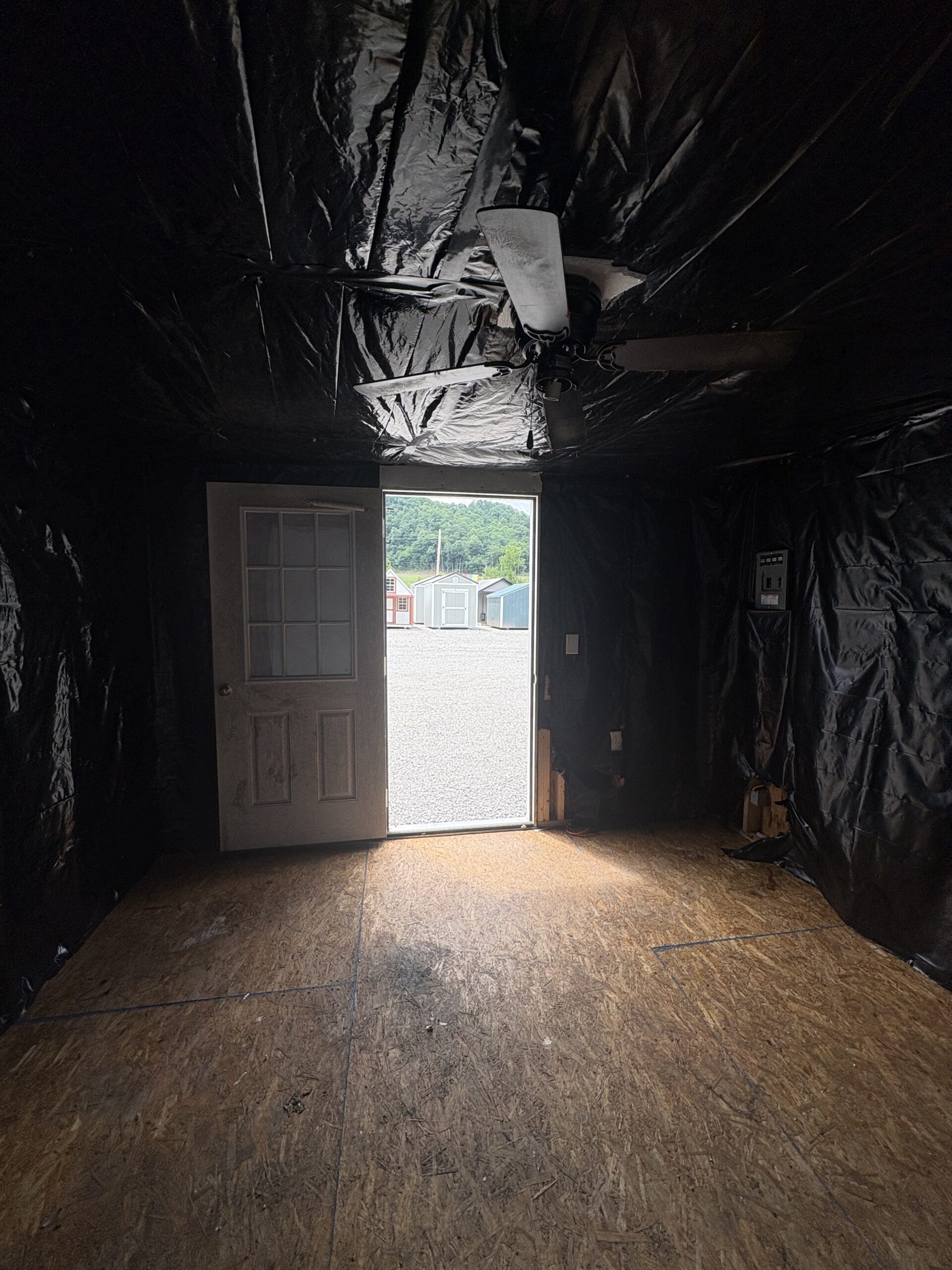 Interior of a dim room lined with black plastic sheeting; open door shows a gravel yard with storage sheds outside and a ceiling fan overhead.