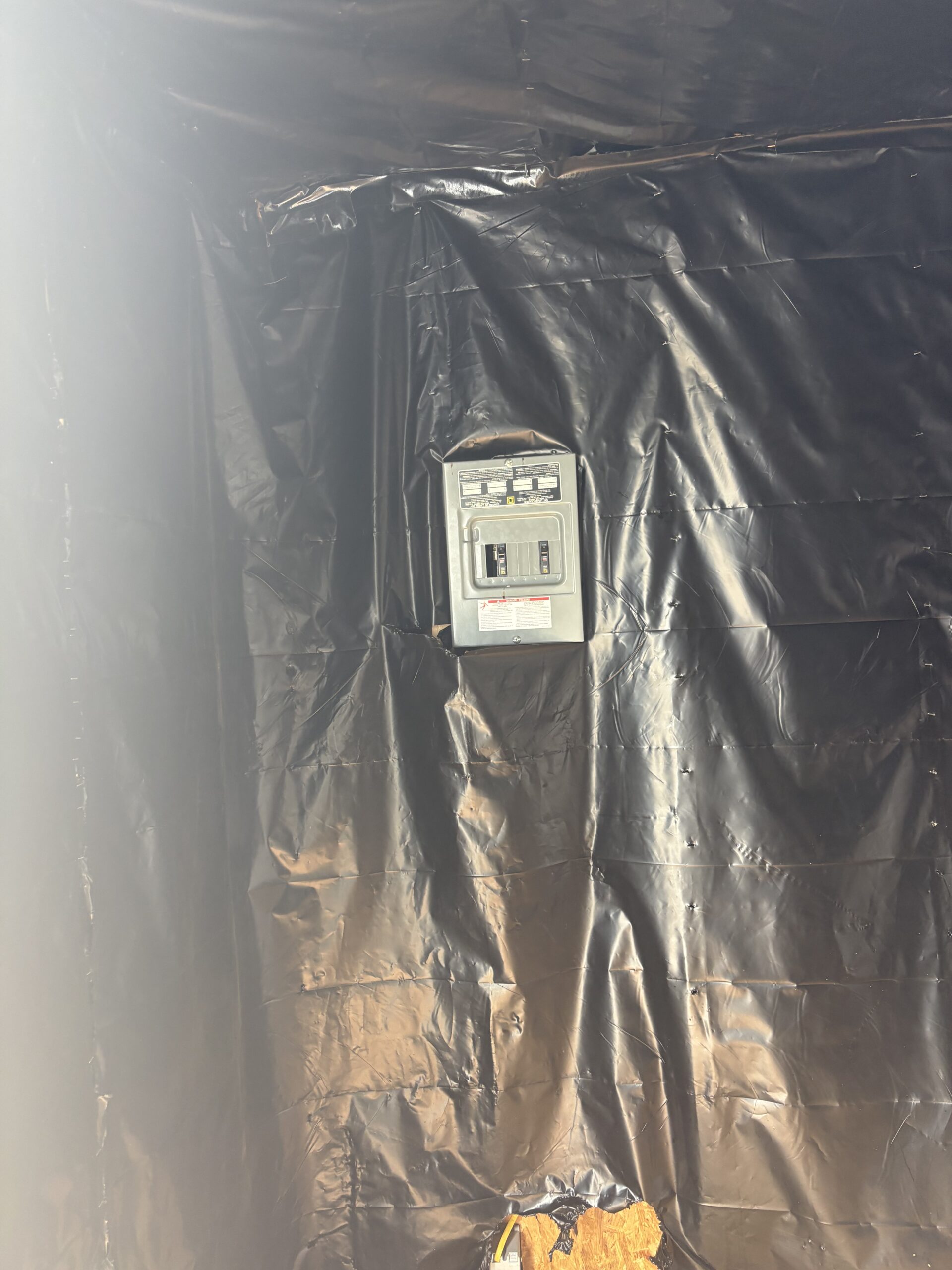 Indoor wall covered in black plastic sheeting with a small electrical breaker panel mounted toward the bottom center; exposed wiring and torn wood nearby.