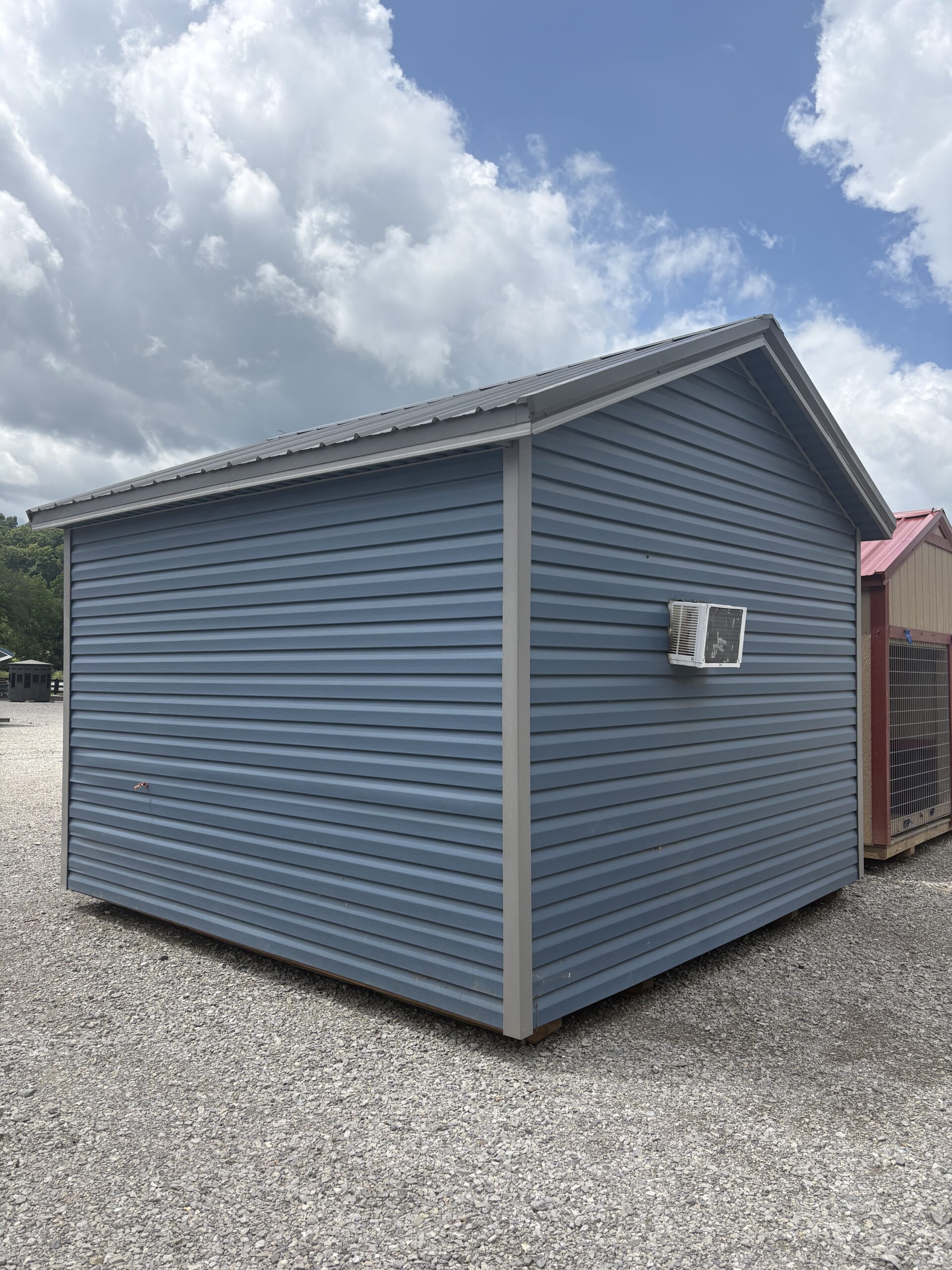 Blue metal shed with white trim and an external air conditioner, on a gravel lot under a cloudy sky.
