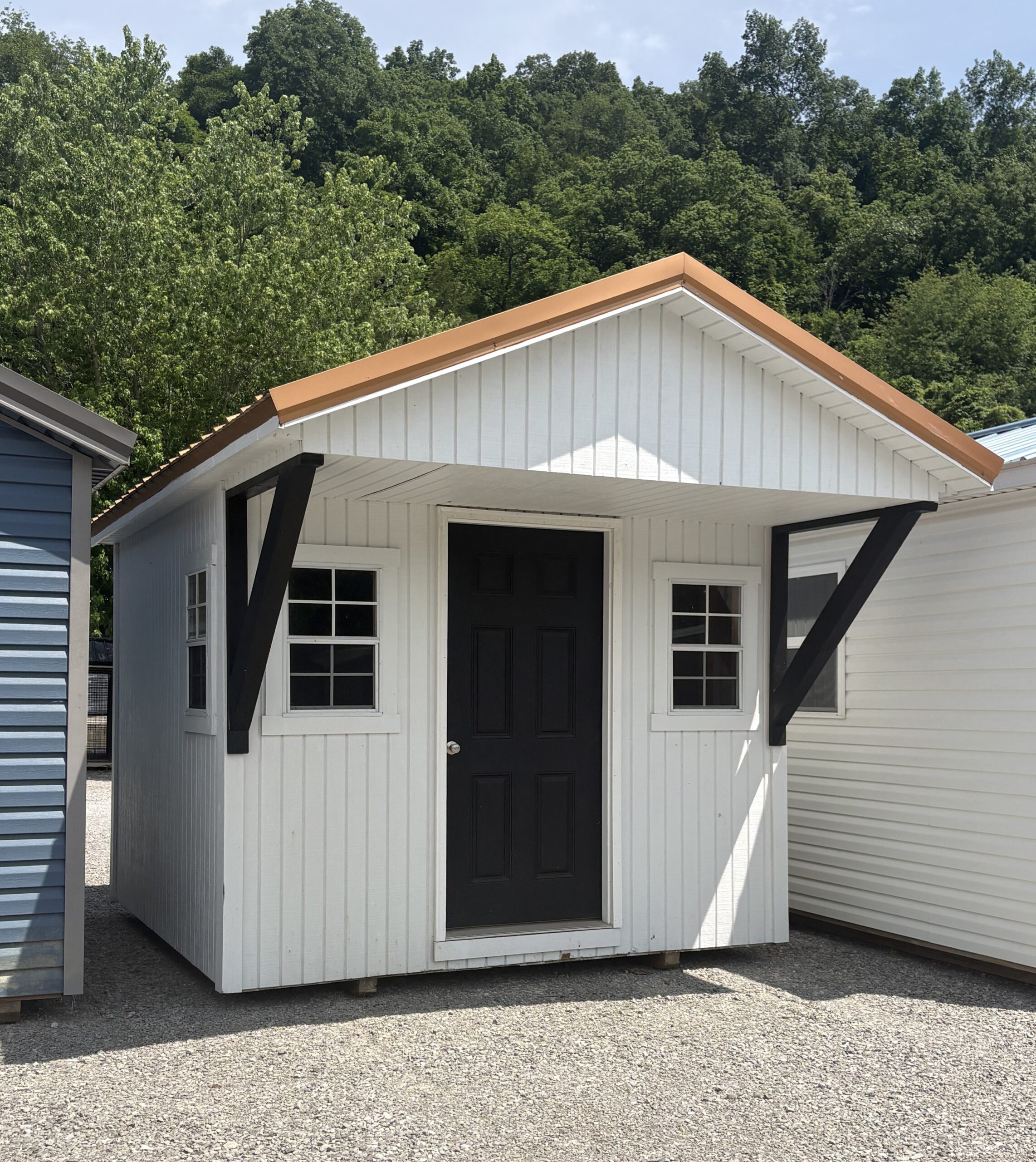 White shed with a black front door, orange trim on the gabled roof, and dark diagonal supports on a gravel lot with trees behind.