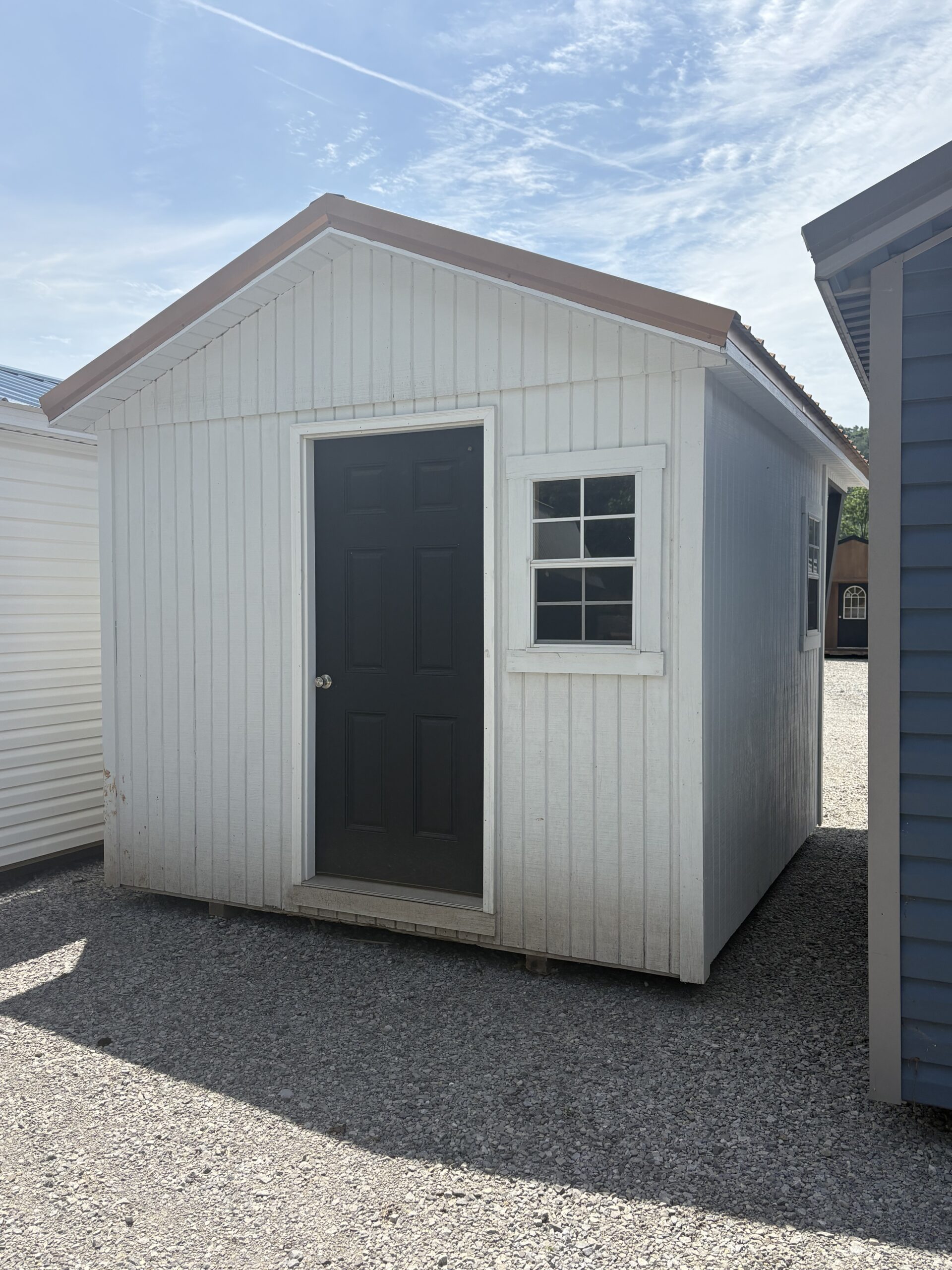 White storage shed with a black front door and a small window, sitting on gravel under a blue sky.