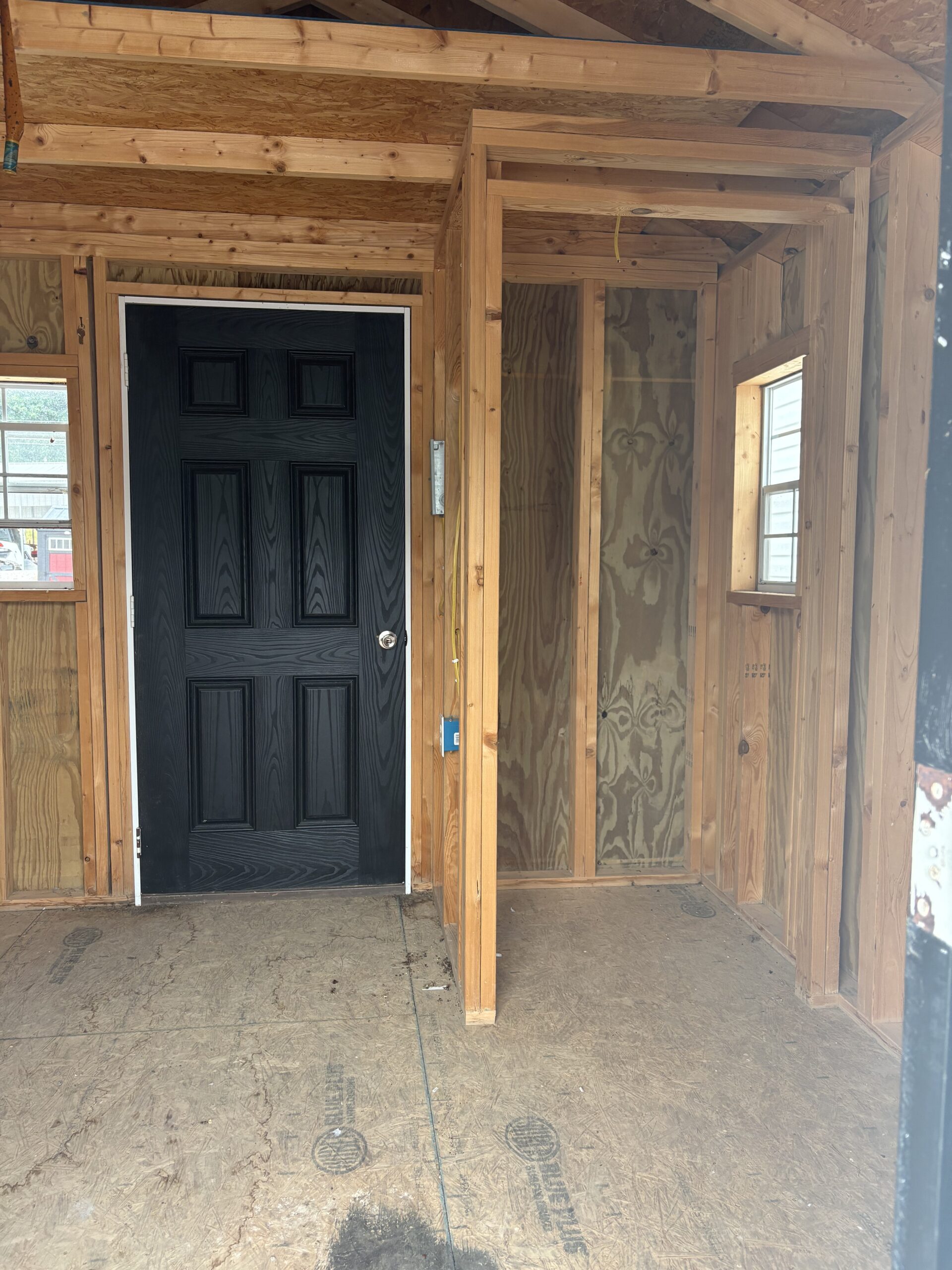 Interior of a partially framed room under construction with a black front door on the left and exposed studs and plywood walls,